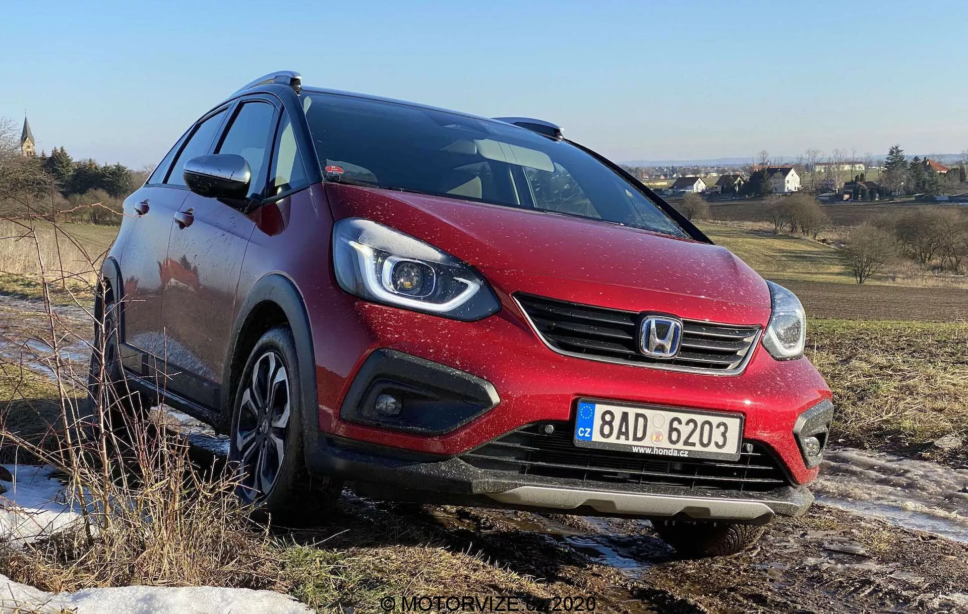 Vista frontal de tres cuartos de un Honda Jazz Crosstar 2020 hatchback rojo, tomada desde un ángulo bajo, mostrando su fascia delantera, faros proyectores de halógeno con luces diurnas LED integradas, parrilla frontal con logotipo de la marca y llantas de aleación. El coche está en un terreno accidentado bajo un cielo azul claro, sugiriendo su capacidad para manejar superficies no pavimentadas.