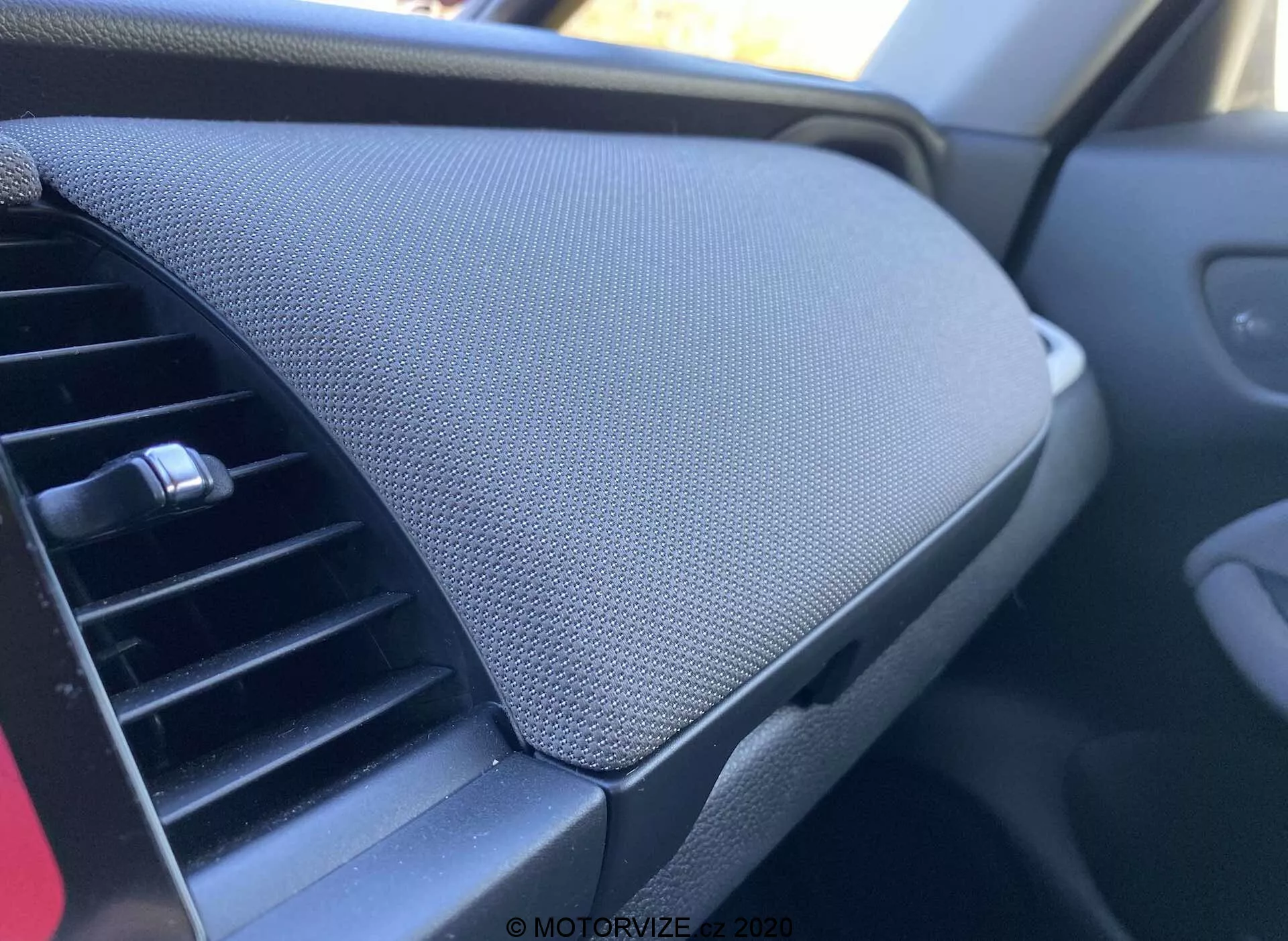 Vista de cerca del interior de un vehículo Honda Jazz 2020 (Jazz Crosstar), centrada en el tablero. Tomada desde la perspectiva del pasajero, la imagen muestra el conducto de aire acondicionado a la izquierda, con un diseño ajustable horizontal. Dominando el lado derecho de la imagen está el compartimiento de almacenamiento del tablero con una tapa cerrada, caracterizada por una superficie texturizada, posiblemente de malla o tela. El compartimiento parece estar integrado sin una manija visible, sugiriendo un mecanismo de apertura a presión. El esquema de color general es oscuro, implicando una estética neutral orientada a reducir distracciones.