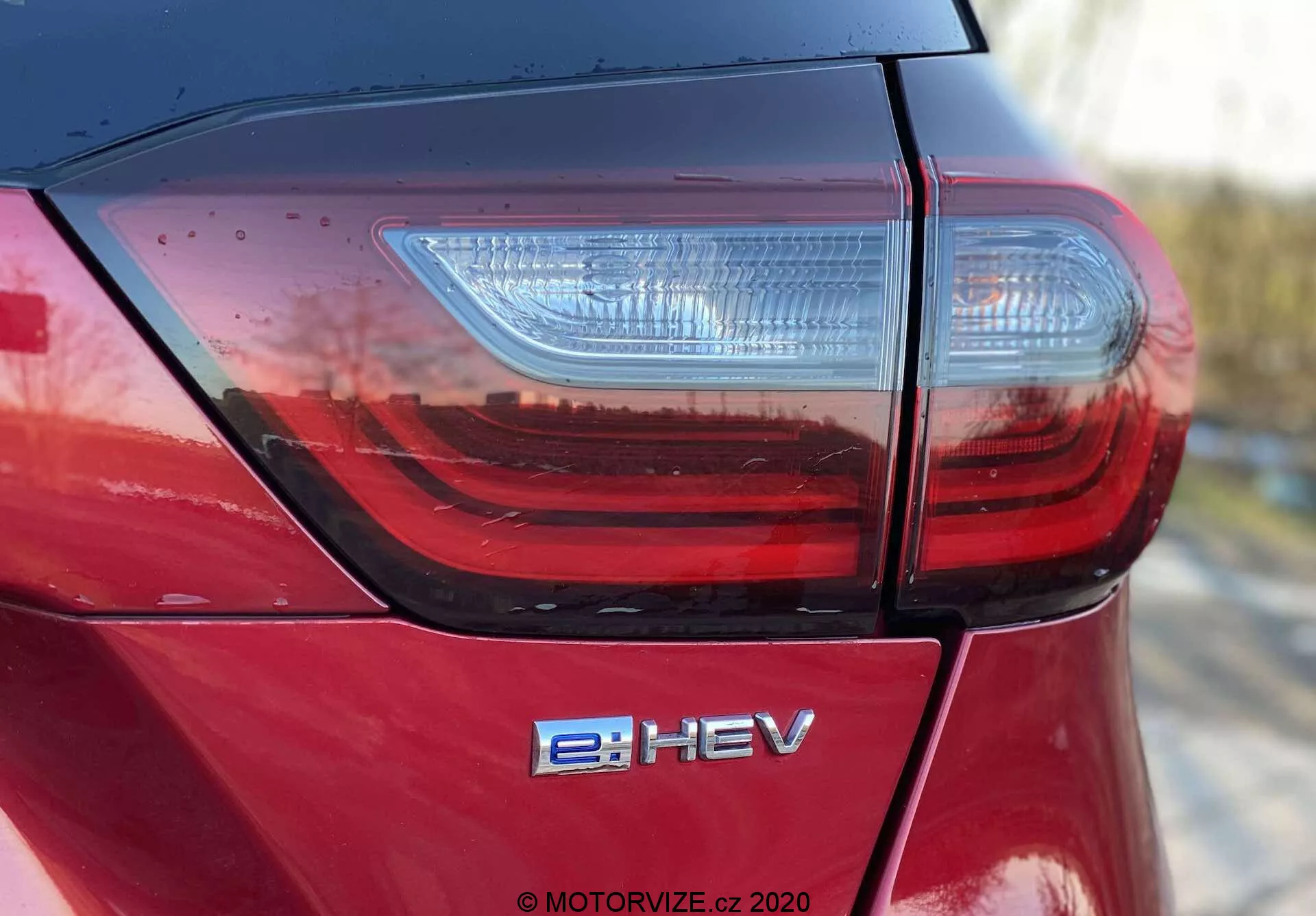 Foto detallada en ángulo alto de la sección trasera de un Honda Jazz Crosstar 2020, que muestra la tapa del maletero, el moderno conjunto de luces traseras LED y la insignia e:HEV de vehículo eléctrico híbrido. Destaca el acabado de pintura metálica roja de alto brillo y la calidad de construcción superior con costuras de paneles ajustadas, indicada por las líneas limpias y los reflejos a la luz natural.