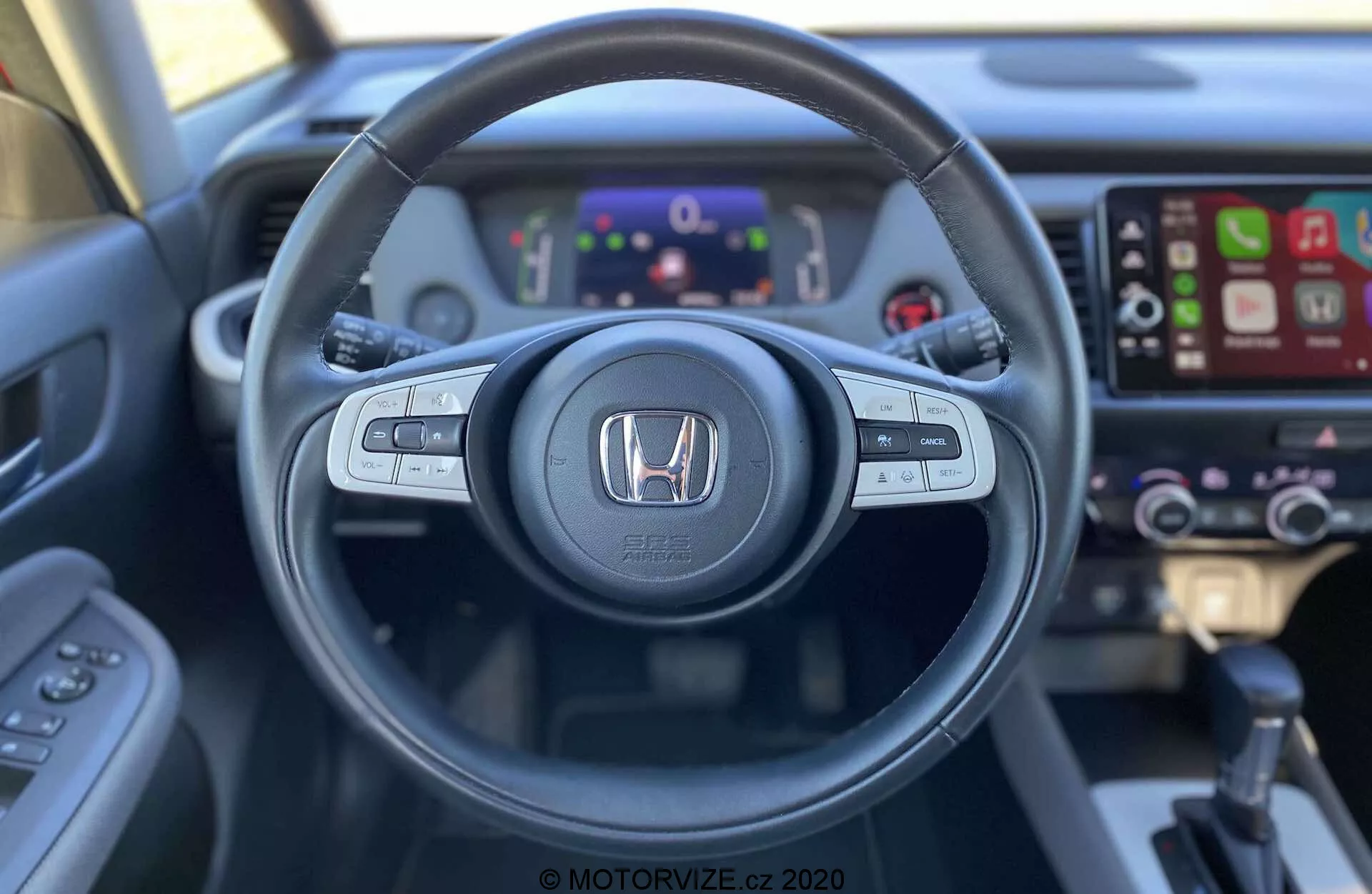 Vista del interior del Honda Jazz Crosstar 2020 desde el asiento del conductor, destacando un volante con botones de control de audio, control por voz y control de crucero. El emblema de Honda se muestra prominente en el centro. Detrás del volante, un cuadro de instrumentos digital muestra varios indicadores e información. A la derecha, se puede ver un sistema de infoentretenimiento con pantalla táctil y compatibilidad con Apple CarPlay, junto con controles de clima ligeramente desenfocados. La imagen, tomada desde una perspectiva de primera persona, resalta el diseño ergonómico y la accesibilidad de la tecnología del vehículo.