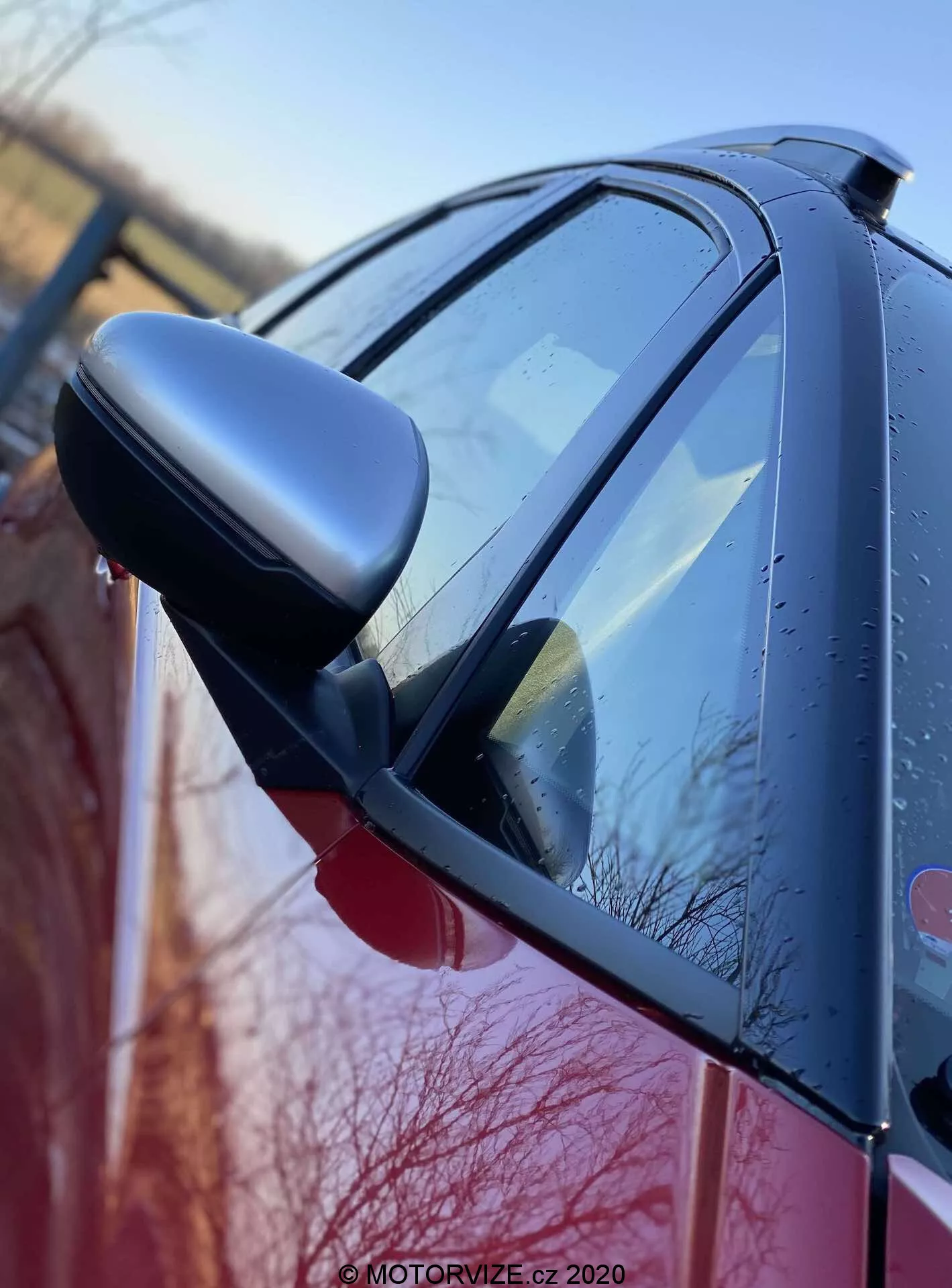 Vista lateral en primer plano del Honda Jazz 2020 (Jazz Crosstar) destacando el espejo retrovisor del lado del conductor con indicadores de señal de giro integrados. La imagen, tomada desde un ángulo bajo cerca de la parte frontal del coche, captura la carrocería brillante, posiblemente roja, con gotas de agua, lo que sugiere lluvia reciente o rocío matutino. El espejo con carcasa oscura y el diseño aerodinámico del coche son características prominentes, enfatizando el acabado y la condición exterior.