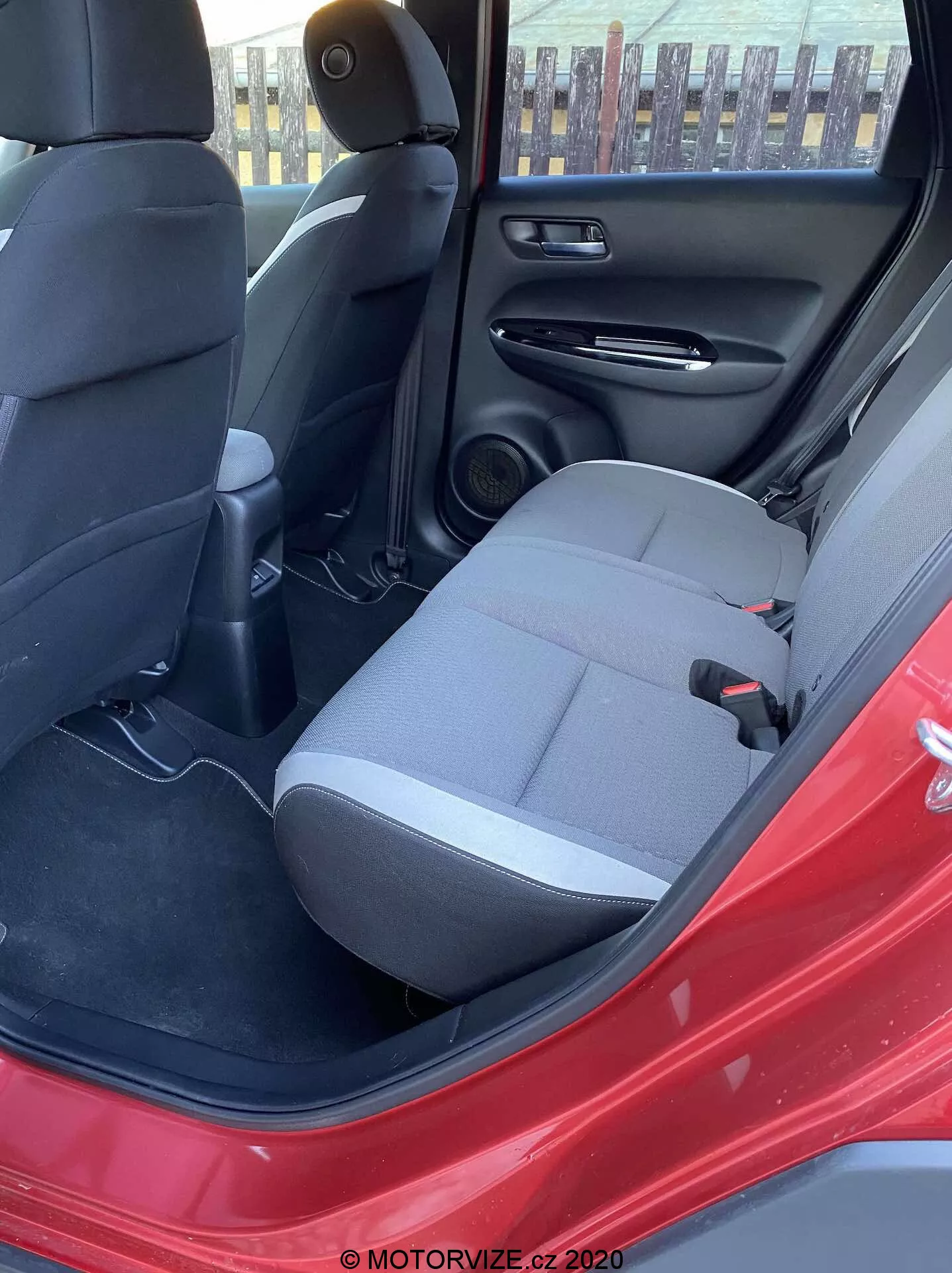 Vista del interior del Honda Jazz Crosstar 2020 desde la puerta trasera del lado del conductor, destacando el asiento trasero del pasajero en tela oscura, cinturón de seguridad, consola central trasera con ventilaciones, altavoz en la puerta trasera, panel de puerta negro con bolsillo de almacenamiento, alfombrado en el piso y el llamativo color rojo del exterior del coche. La luz natural diurna ilumina la escena.