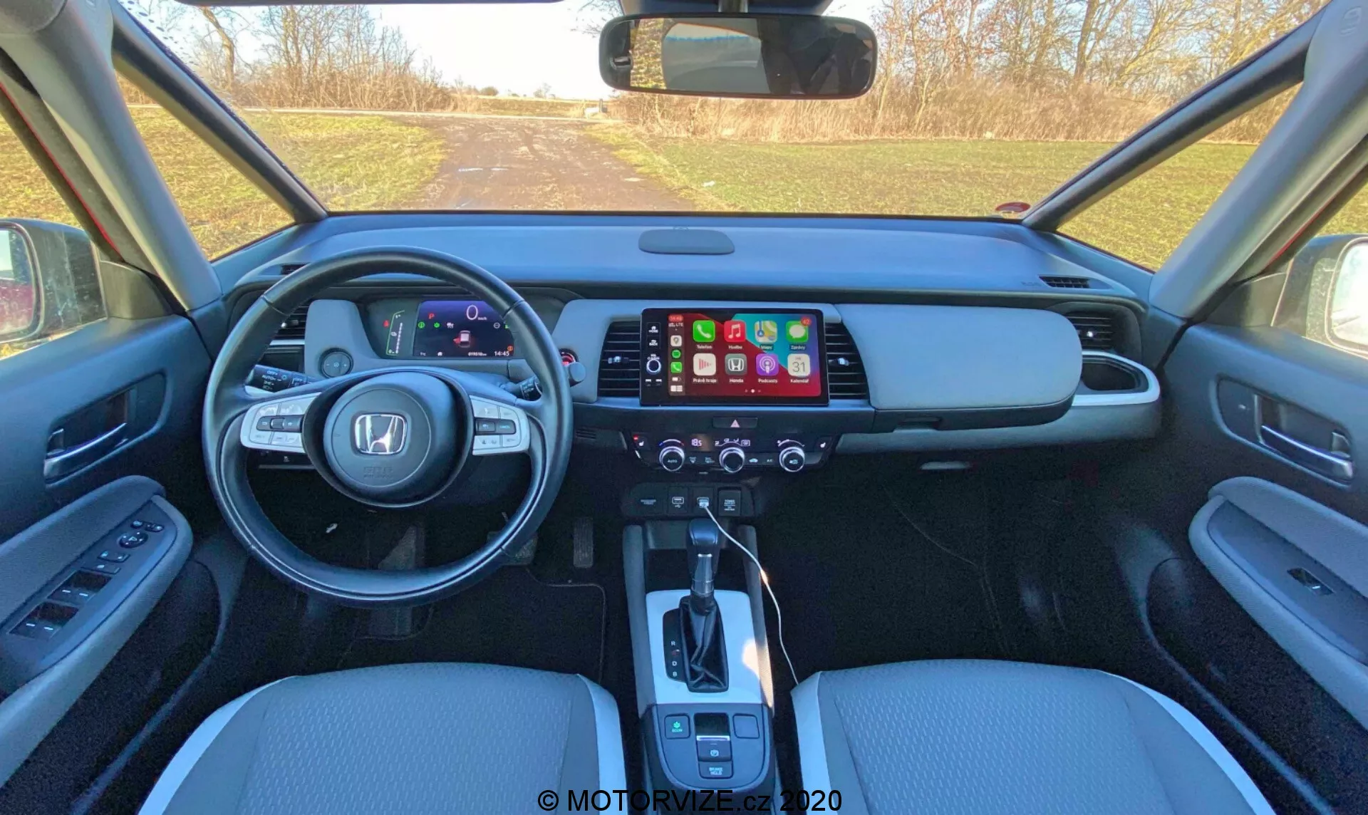 Vista del interior del Honda Jazz 2020 (Jazz Crosstar) desde la zona de asientos traseros, mostrando el tablero frontal con un panel de instrumentos digital, una gran pantalla táctil de infoentretenimiento, panel de control del clima, palanca de cambios automática, asientos de cuero y controles del panel de la puerta.