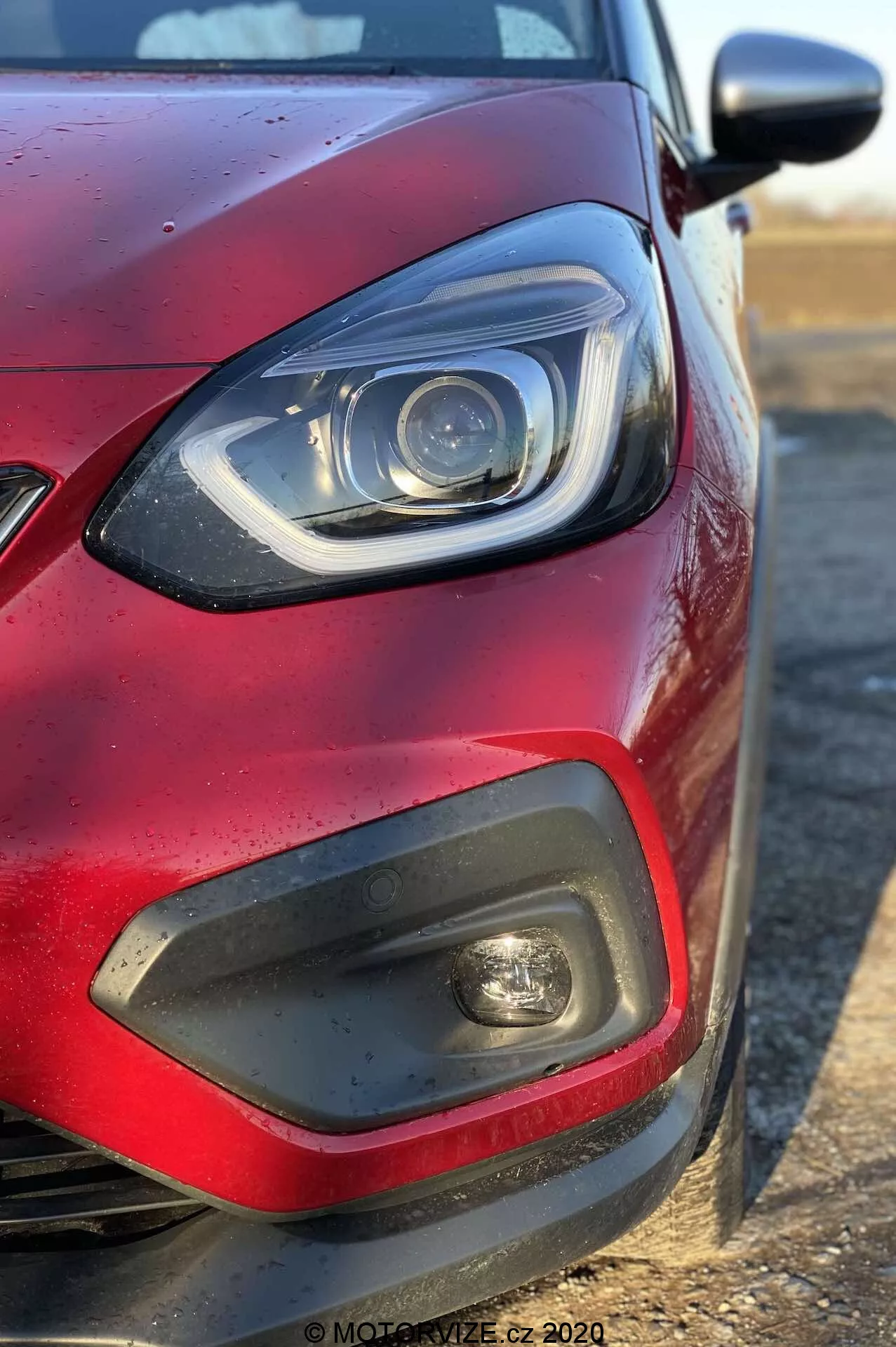 Primer plano del cuarto delantero izquierdo de un Honda Jazz 2020 (Jazz-Crosstar) rojo desde un ángulo lateral, enfocado en el faro con elementos LED o de xenón y una barra DRL curvada, un inserto de plástico negro con luz antiniebla, una parte de la rejilla, pintura roja metálica con gotas de agua y un espejo lateral del color de la carrocería que se extiende desde la derecha.