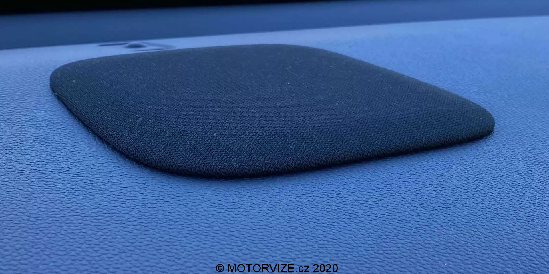 Fotografía detallada de una rejilla de altavoz de coche incrustada en el panel del salpicadero o puerta de color azul oscuro o negro, posiblemente del Honda Jazz 2020. La rejilla del altavoz está hecha de un material texturizado similar a la tela y tiene forma de trapezoide truncado o cuadrilátero irregular con un lado ligeramente convexo. El componente está ligeramente elevado, proyectando una sombra que enfatiza su perfil y textura. La foto está tomada desde un ángulo lateral utilizando luz natural difusa, destacando la textura granulada de la superficie circundante y contiene una marca de agua que dice '© MOTORVIZE.cz 2020.'