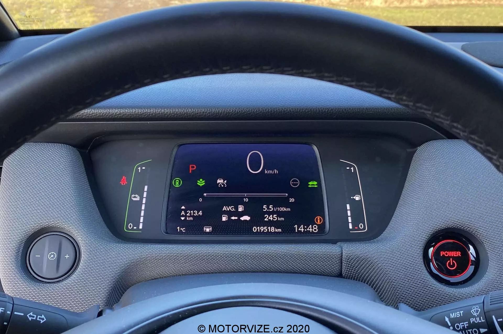 Primer plano del interior del Honda Jazz Crosstar 2020 desde el asiento del conductor, mostrando el volante y el cuadro de instrumentos totalmente digital. El volante envuelto en tela obstruye parcialmente la vista de la pantalla digital, que muestra símbolos del sistema del coche, un gráfico circular central, información del viaje, odómetro y reloj. Dos palancas detrás del volante controlan los intermitentes, las luces y los limpiaparabrisas. Un panel de control para los faros y un botón de encendido con un anillo rojo son visibles a la izquierda y derecha, respectivamente. Los materiales del tablero parecen texturizados, destacando un diseño ecológico. La imagen enfatiza la tecnología moderna, la instrumentación digital y las posibles características de vehículos híbridos o eléctricos.