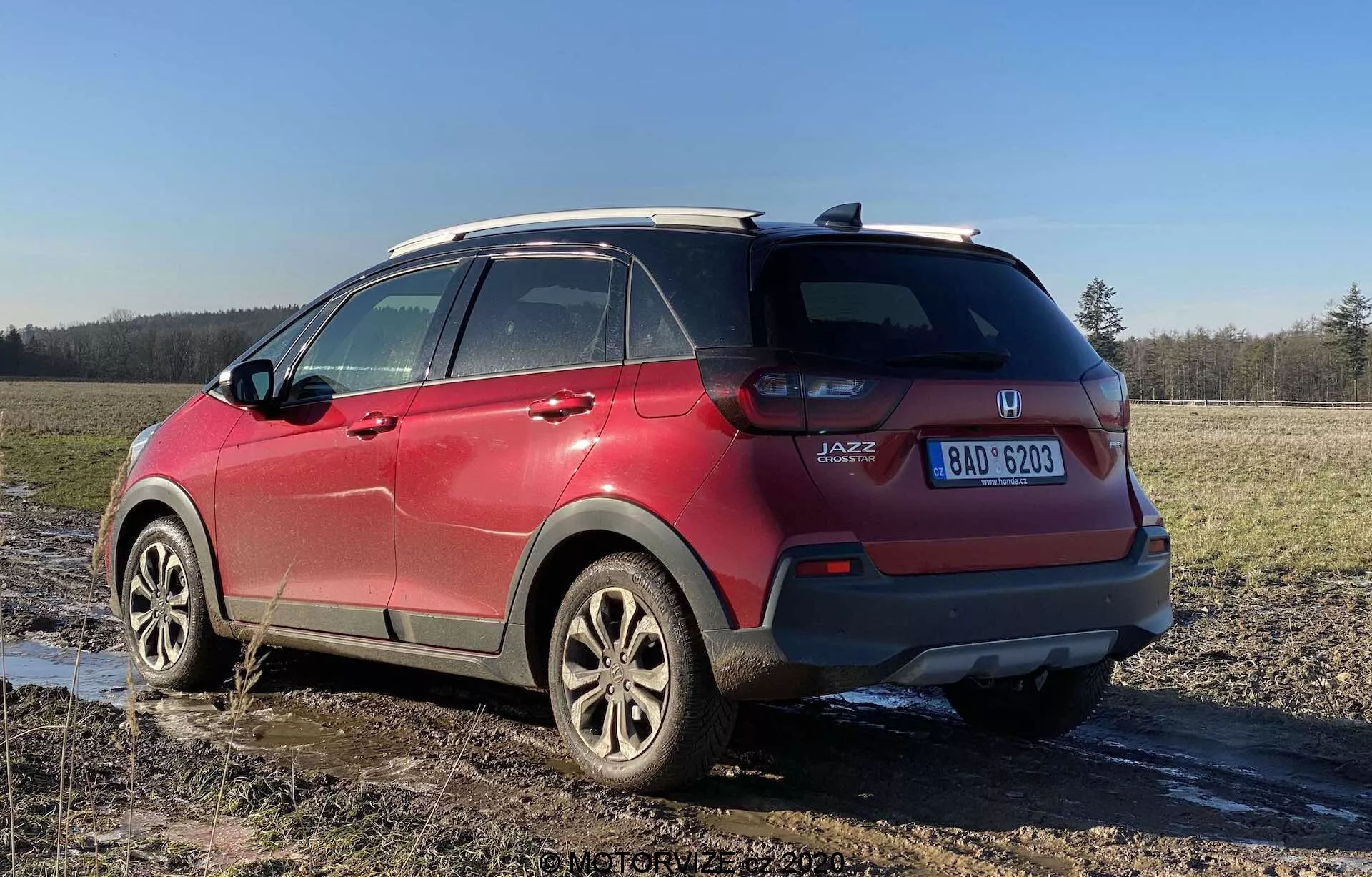 Vista trasera en cuarto de un Honda Jazz Crosstar 2020 rojo, destacando sus características de SUV crossover subcompacto. La imagen capturada desde un ángulo bajo resalta la altura y la distancia al suelo del vehículo, con clusters de luces traseras visibles, insignia 'JAZZ', llantas de aleación, sistema de rieles en el techo, ventanas traseras tintadas, salpicaduras de barro y una placa de matrícula visible, estacionado en un campo embarrado.