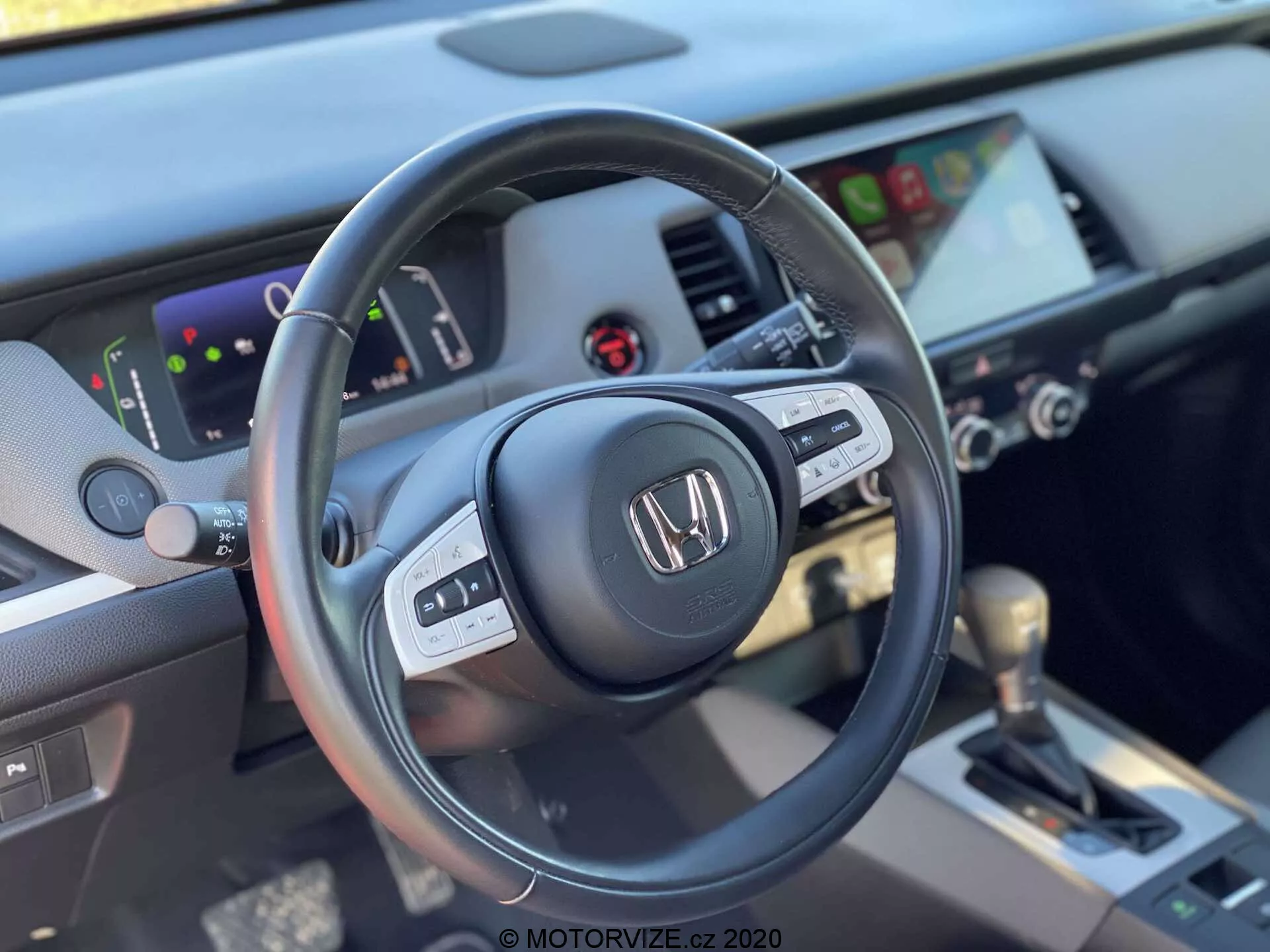 Vista de cerca del interior del Honda Jazz Crosstar 2020 tomada desde el asiento del conductor, resaltando el lado izquierdo del volante de tres radios forrado en cuero con controles multifuncionales y el tablero con un clúster de instrumentos digital y un sistema de infoentretenimiento táctil. La imagen incluye controles HVAC y una palanca de cambios automática forrada en cuero en la consola central, enfatizando un diseño moderno y bien equipado.