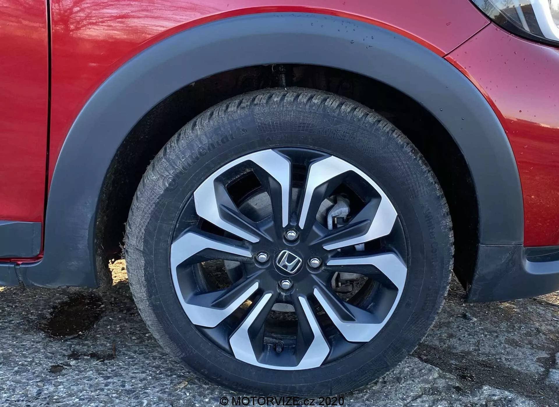 Toma de primer plano de la rueda delantera izquierda y la carrocería inferior del Honda Jazz Crosstar 2020, destacando la moderna rueda de aleación de múltiples radios con un acabado bicolor en plata y negro, el diseño de la banda de rodadura y la información del flanco del neumático. La imagen también muestra el arco del guardabarros, parte del parachoques delantero y una sección de la puerta lateral desde un ángulo ligeramente bajo, resaltando aspectos de diseño y tecnología, como la tecnología de neumáticos, el diseño de ruedas, la composición del material, la aerodinámica y la configuración de la suspensión del vehículo.