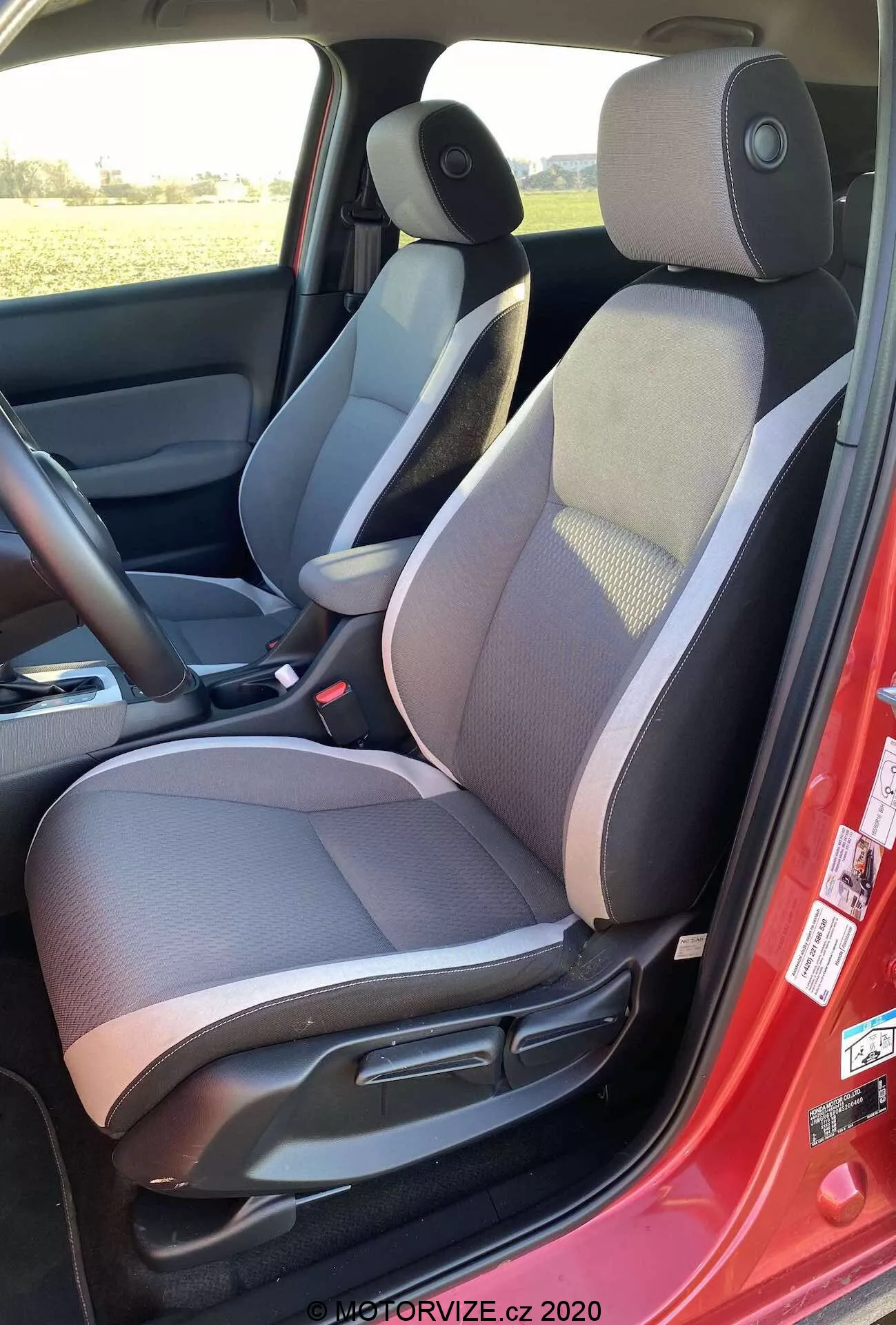 Vista interior del Honda Jazz Crosstar 2020 desde el lado izquierdo, destacando el asiento del conductor con tela de dos tonos y sección central con diseño, reposacabezas ajustable, controles de ajuste manual del asiento, cinturón de seguridad rojo, altavoz en el panel de la puerta, alfombra oscura, consola central con reposabrazos, control del espejo lateral y esquema de colores oscuro y claro.