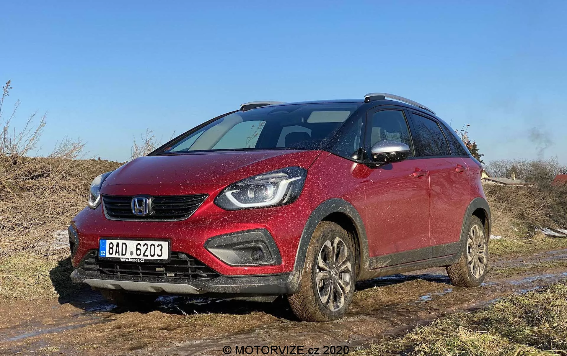 Vista en ángulo frontal de un Honda Jazz Crosstar 2020 rojo, un SUV crossover compacto, capturado en un terreno todoterreno con tierra y barro visibles. La imagen destaca la parrilla delantera, los faros elegantes que posiblemente albergan lámparas LED o de proyector, los faros antiniebla integrados en el parachoques y un capó suavemente inclinado. El vehículo, con barras de techo, ruedas cubiertas de barro y espejos laterales con señales de giro integradas, sugiere características modernas de seguridad y equipo tecnológico avanzado. Ideal para categorías centradas en vehículos, reseñas de autos, capacidades todoterreno, tecnología automotriz y diseño.