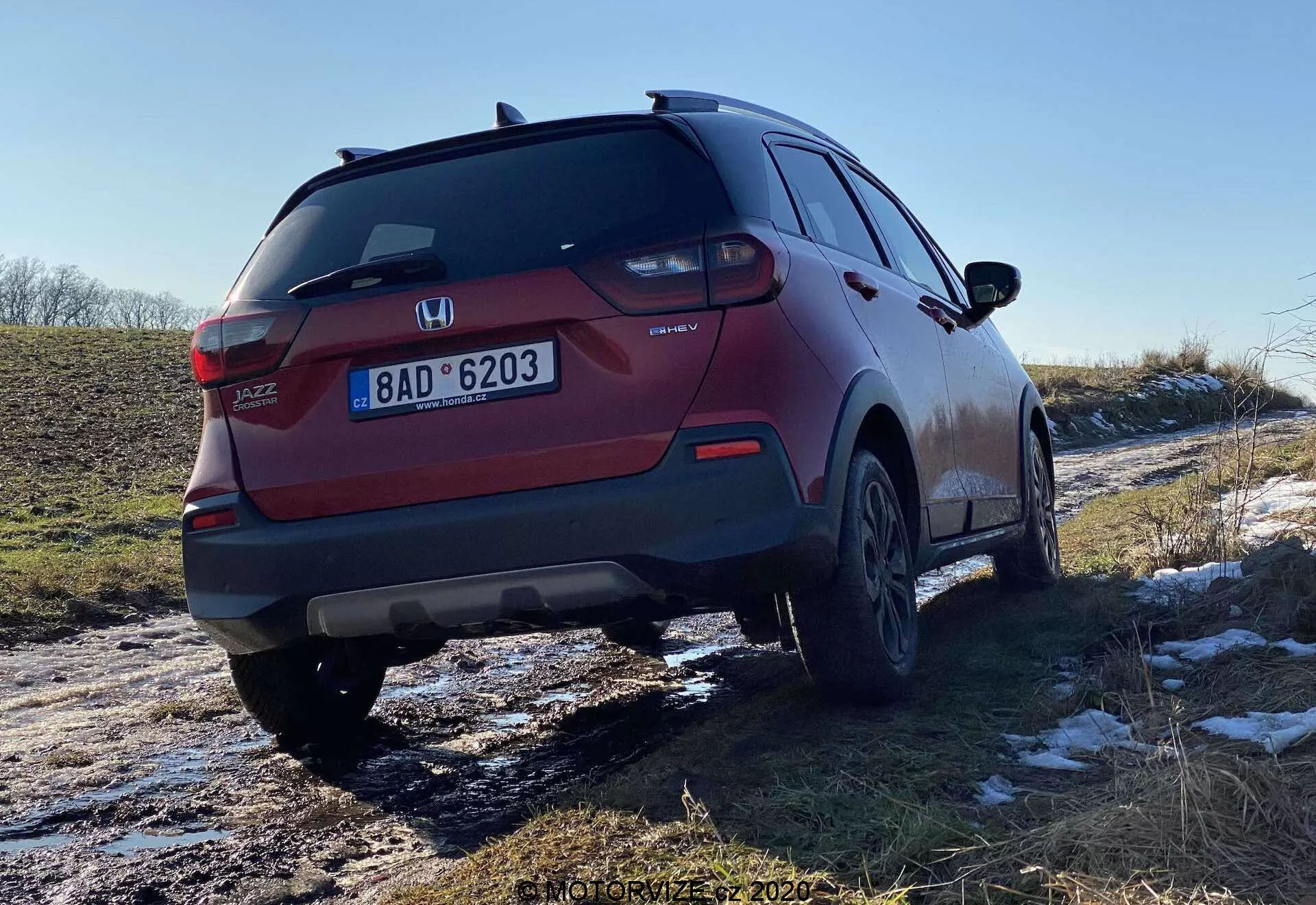 Vista trasera de tres cuartos de un SUV Honda Jazz Crosstar rojo, que muestra el diseño de las luces traseras, el parachoques trasero, el sistema de escape y las ruedas de aleación. El ángulo enfatiza la distancia al suelo del vehículo y parece haber viajado recientemente por carreteras nevadas y embarradas, lo que indica su versatilidad en varias condiciones.