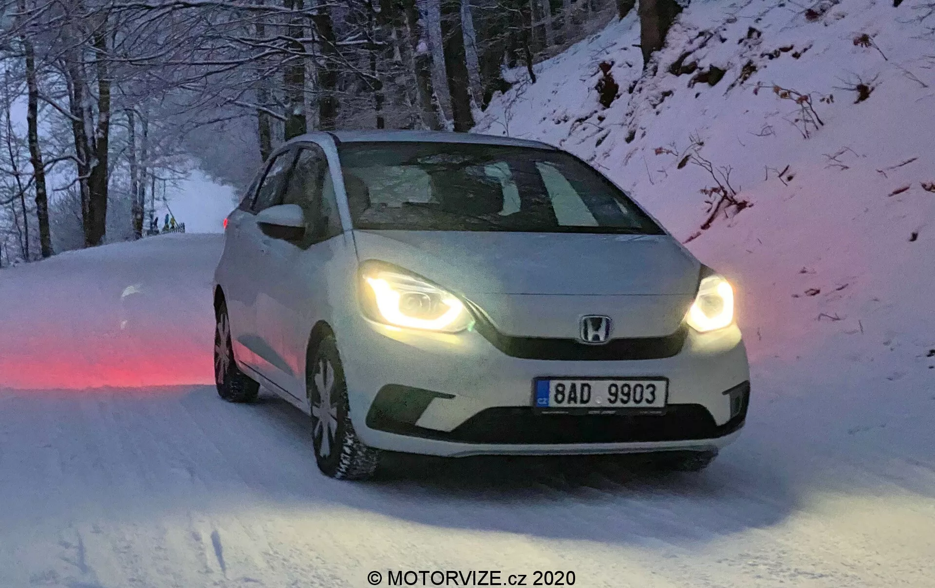 Esta imagen muestra un Honda Jazz 2020, capturado desde un ángulo de tres cuartos frontales, exhibiendo las características de diseño de la parrilla frontal, los faros y los contornos del capó. Posicionado en un camino cubierto de nieve, el vehículo tiene neumáticos de invierno con patrones de banda de rodadura visibles y los faros encendidos, indicando condiciones de amanecer, anochecer o baja visibilidad. La placa europea dice “BAD 9903”, y la imagen tiene una ligera borrosidad de movimiento que añade una cualidad dinámica. La iluminación natural y fría y el entorno invernal se complementan con las capacidades para todo tipo de clima del automóvil.
