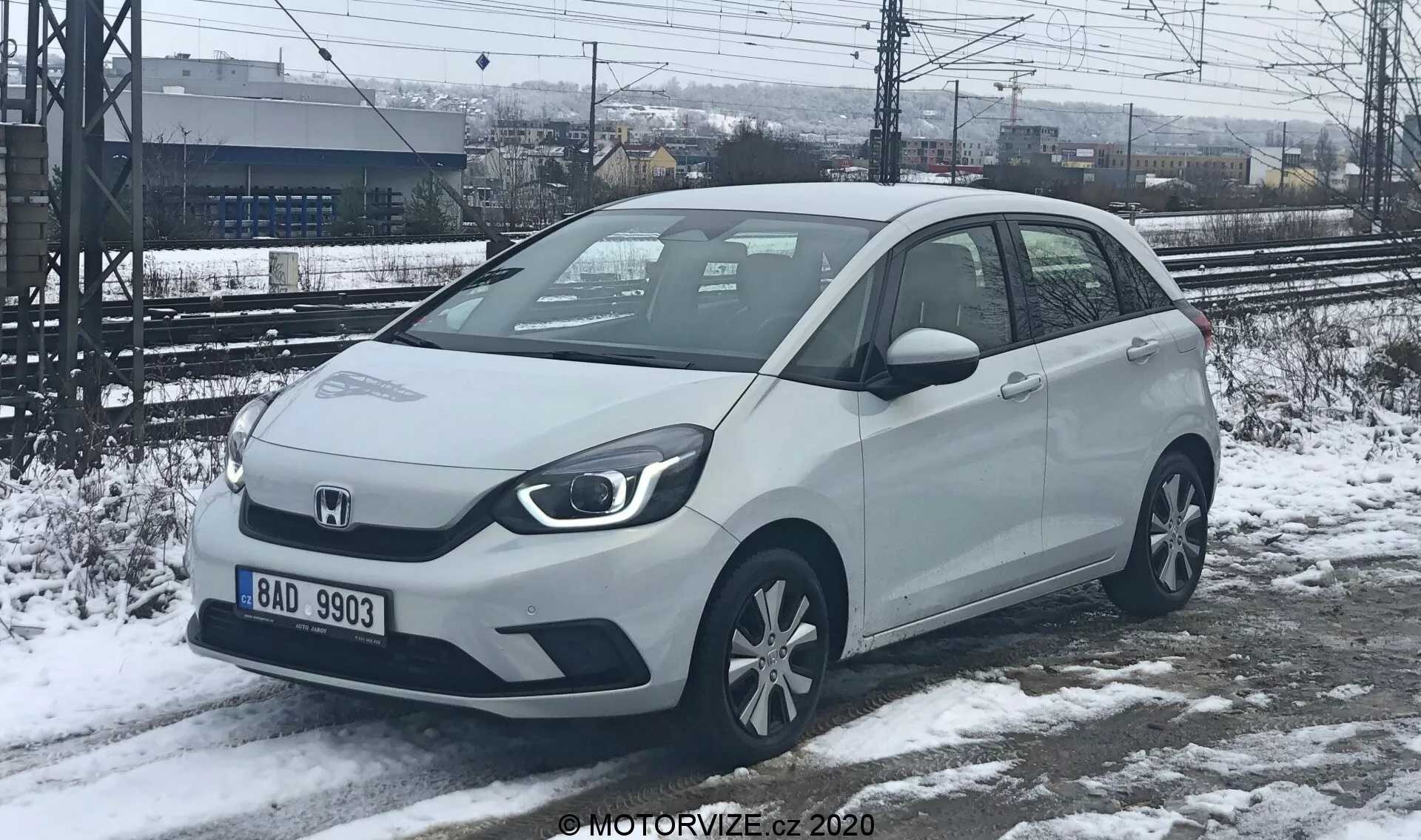 Vista en ángulo de tres cuartos delantero de un coche compacto Honda Jazz 2020 de color claro, posiblemente blanco o plateado, capturado en un entorno invernal con nieve en el suelo. Los faros angulares y la parrilla delantera sólida sugieren opciones de tren motriz electrificado. El coche tiene sensores de aparcamiento visibles en el parachoques delantero, espejos laterales montados en las puertas con señales de giro integradas y llantas de aleación estándar. Aparcado en nieve parcialmente derretida, con vías de tren y una cerca de fondo, indicando una ubicación industrial o una estación de tren.