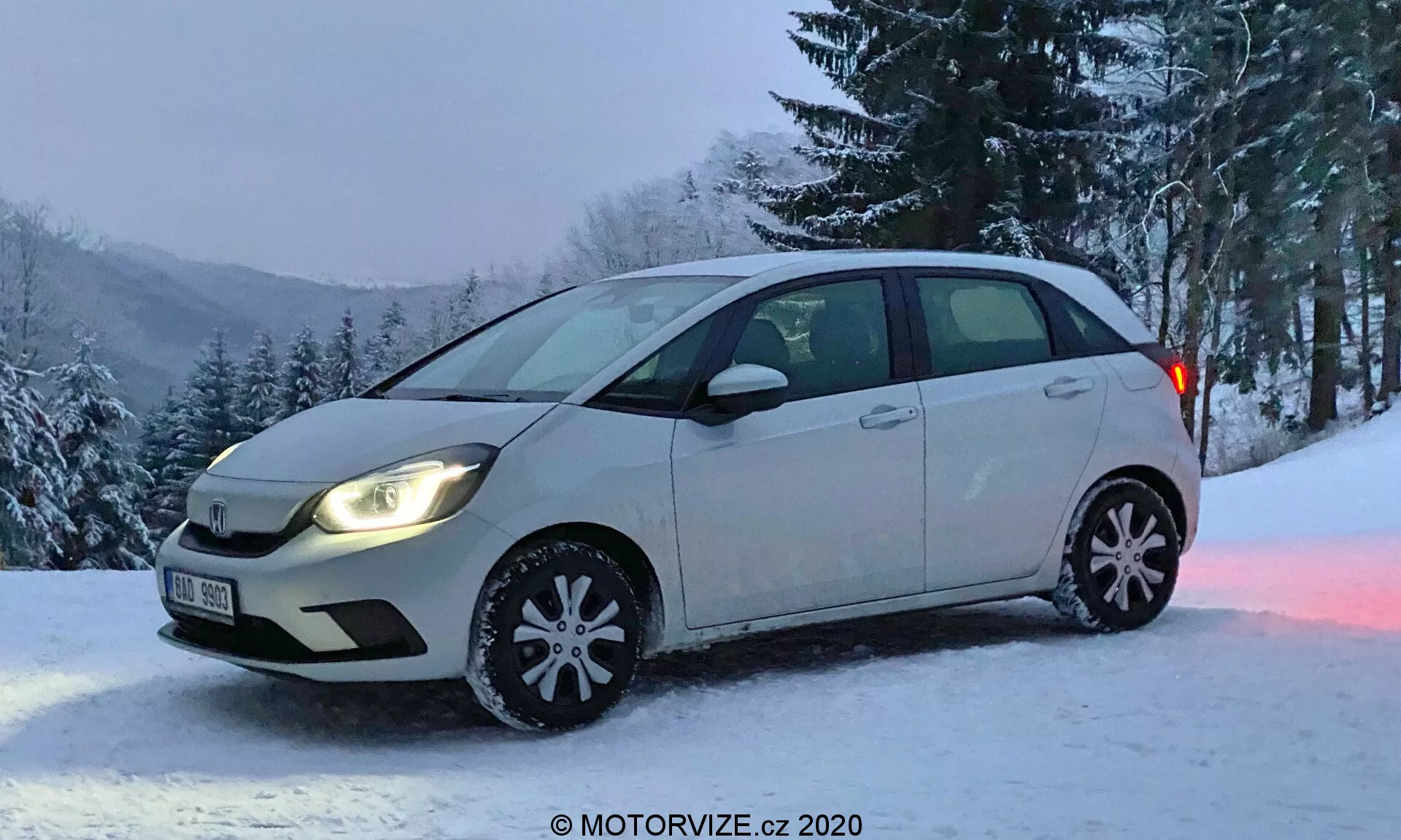 Honda Jazz 2020 hatchback compacto fotografiado desde un ángulo frontal oblicuo con vista completa del lado del conductor y vista parcial del parachoques y la parrilla delanteros. El vehículo presenta un esquema de color de carrocería de dos tonos, con la mayor parte en un color más claro y la parte inferior de las puertas laterales en un tono más oscuro. Las luces delanteras están encendidas, con bombillas halógenas típicas, sin luces antiniebla. Las cubiertas decorativas de las ruedas de invierno son visibles con copos de nieve reflectantes, indicando una temporada de invierno. El coche está estacionado en una zona nevada con un fondo de árboles y una colina nevada, lo que sugiere una ubicación rural o montañosa. Imagen cortesía de MOTORVIZE.cz.