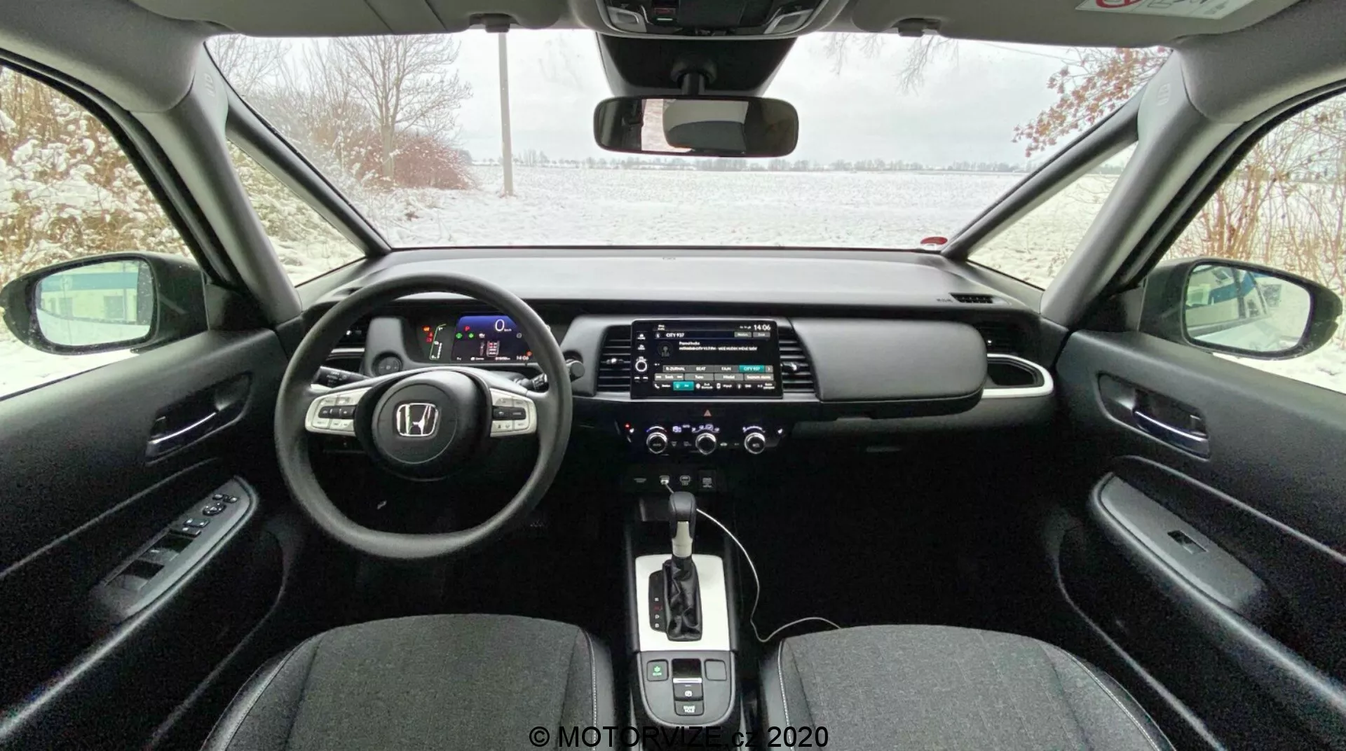 Interior moderno de automóvil visto desde el asiento trasero, que incluye un cuadro de instrumentos digital, volante multifuncional, gran pantalla táctil de infoentretenimiento, caja de cambios automática con palanca de cambio cubierta de cuero, control de clima de doble zona, freno de mano electrónico y diseño elegante con materiales contrastantes; exterior nevado a través del parabrisas, lo que indica una posible ambientación invernal.