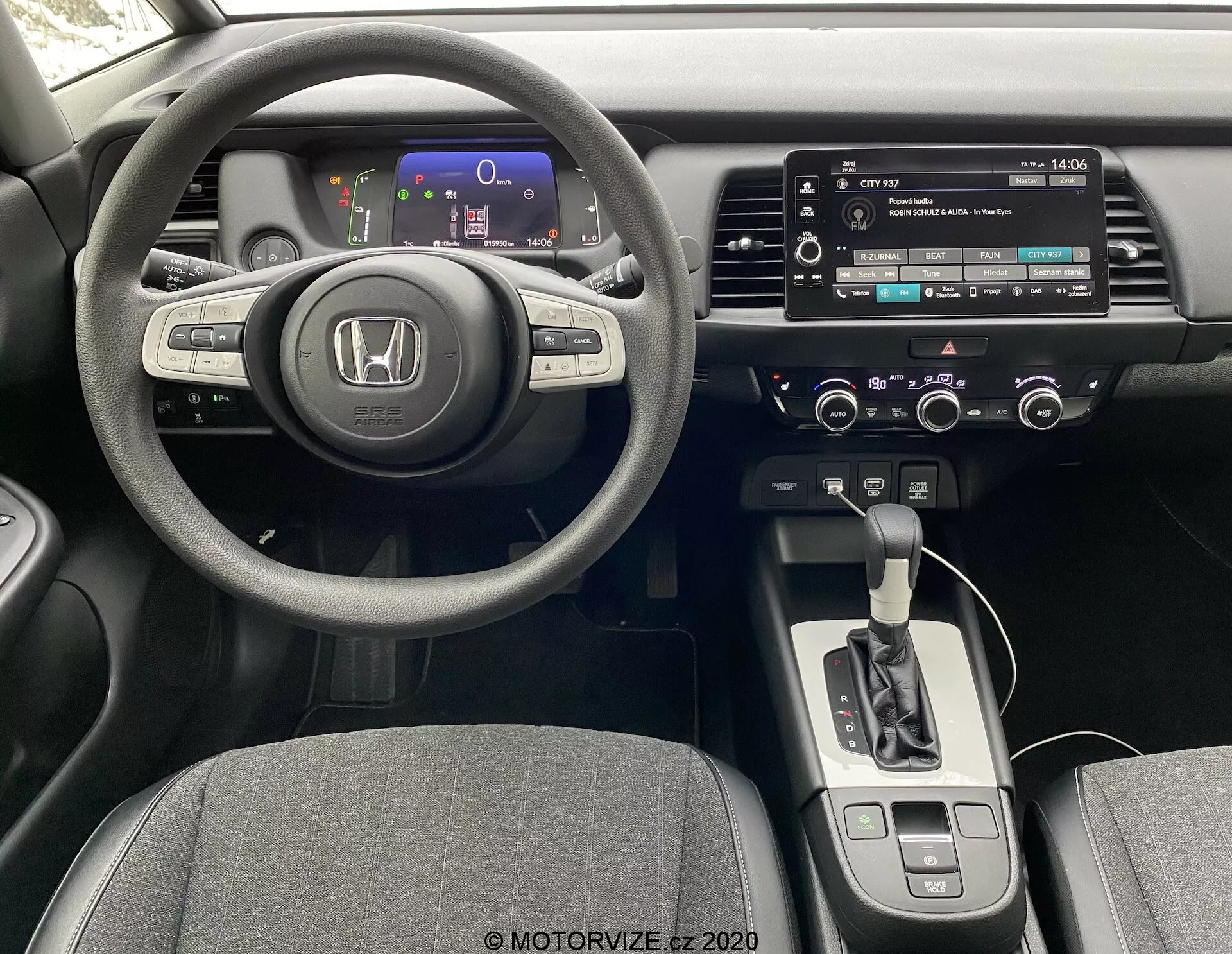 Interior de un Honda Jazz 2020 desde la perspectiva del conductor, mostrando un volante de tres radios con controles integrados, un cuadro de instrumentos digital con indicadores de colores, una consola central con sistema de infoentretenimiento con pantalla táctil, controles manuales de audio y clima, un selector de la transmisión automática, y asientos tapizados en tela.