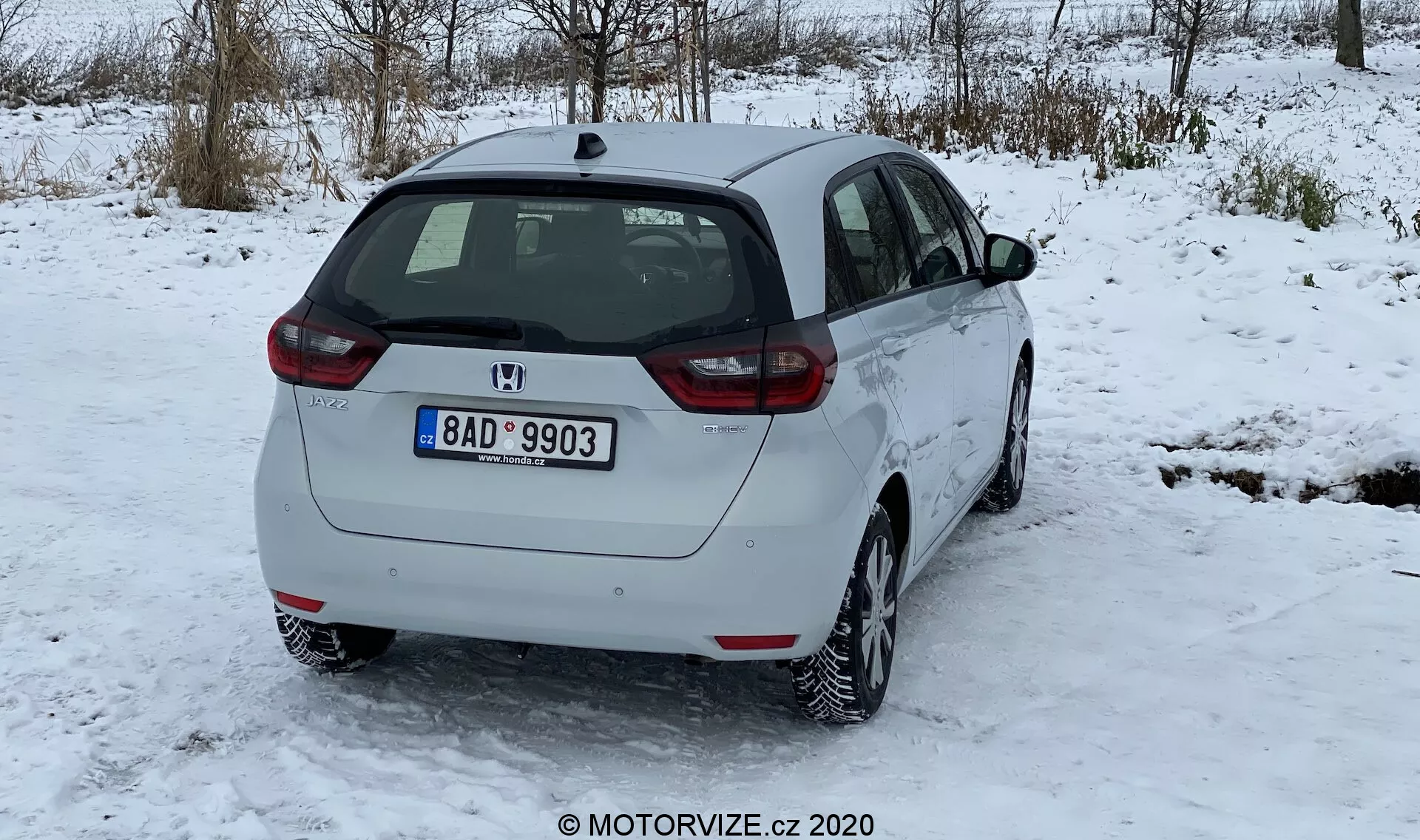 La imagen muestra una vista tres cuartos trasera de un Honda Jazz 2020 blanco, estacionado sobre una superficie cubierta de nieve, con árboles sin hojas en el fondo de un paisaje invernal y un cielo nublado. Las características del diseño trasero incluyen un limpiaparabrisas trasero, el emblema del fabricante, la indicación del modelo y la asamblea de iluminación trasera con lentes rojas y claras. El perfil lateral muestra la puerta trasera de pasajeros, la puerta delantera, las ventanas laterales y un tapón de combustible visible. El coche está equipado con ruedas de aleación de múltiples radios, indicando un dibujo de neumáticos adecuado para nieve con una acumulación menor de nieve. La matrícula trasera sugiere su origen geográfico. La distancia al suelo es típica de los hatchbacks, sin punta de escape visible ni accesorios exteriores. El coche parece limpio y en buen estado, con poca suciedad en el parachoques trasero debido a carreteras nevadas.