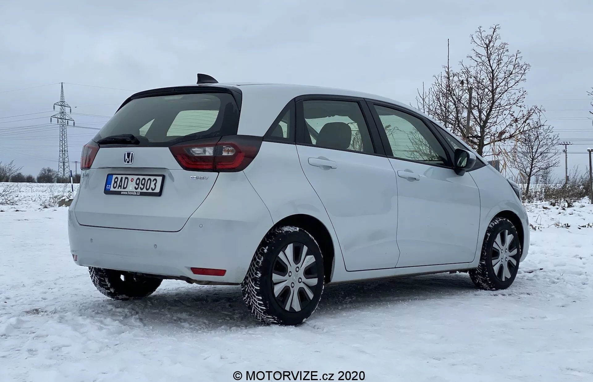 Vista tres cuartos trasera de un coche hatchback Honda Jazz 2020 sobre una superficie cubierta de nieve en un entorno suburbano con árboles desnudos y torres eléctricas en el fondo. La imagen captura el lado del pasajero del coche, el diseño moderno de las luces traseras probablemente con tecnología LED, la puerta trasera, el limpiador del parabrisas trasero, el paragolpes trasero visible con sensores de aparcamiento integrados y neumáticos de invierno aparentes. El vehículo está estacionado bajo luz natural con un cielo nublado, sugiriendo condiciones de conducción invernales.
