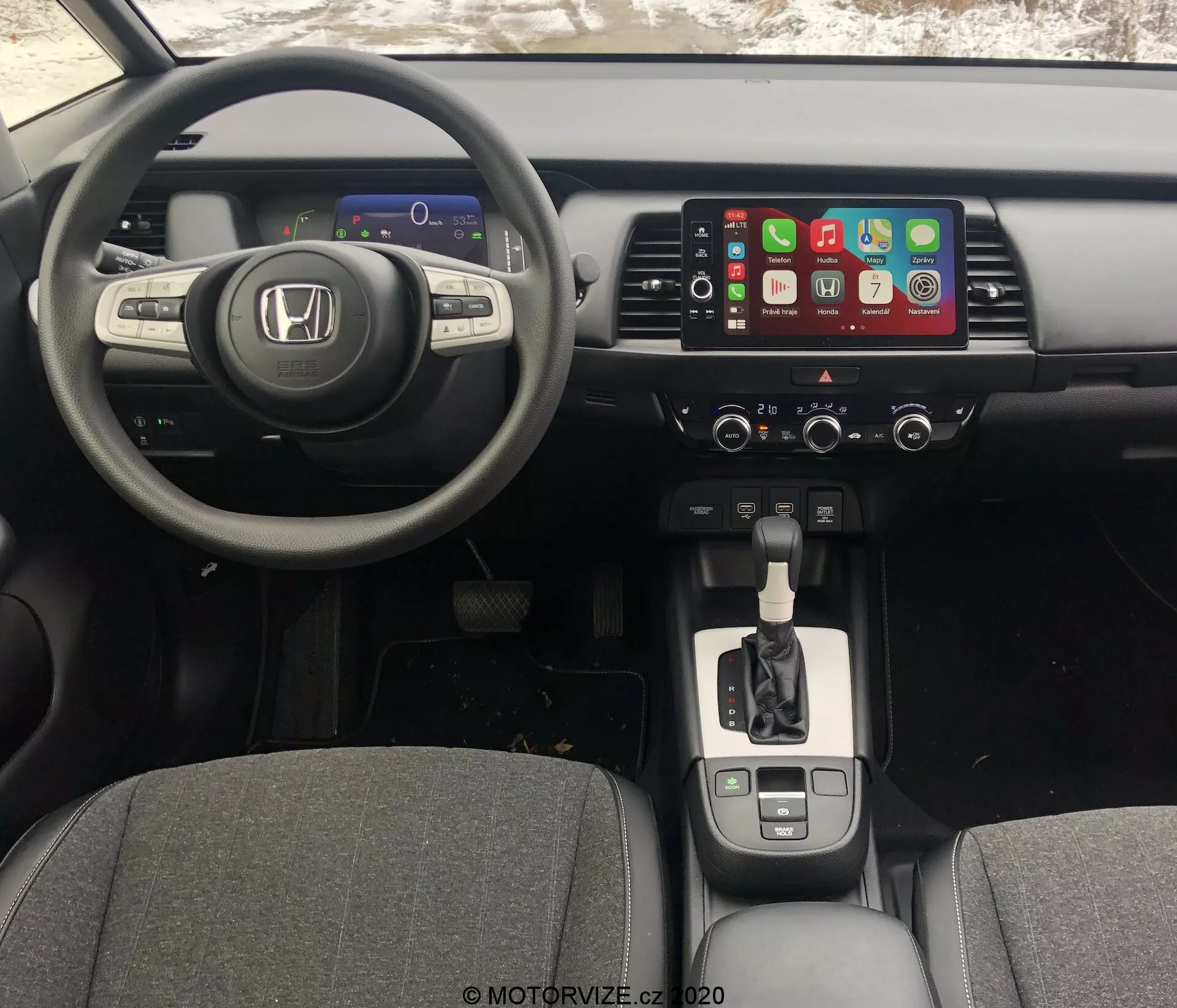 Vista interior del Honda Jazz 2020 desde la perspectiva del conductor, mostrando el tablero, el volante y la consola central. El ángulo ligeramente elevado revela el panel de instrumentos digital, el volante multifuncional de tres radios, el sistema de infoentretenimiento táctil con Apple CarPlay, los controles del clima, la palanca de cambios con acabado cromado, los portavasos, el freno de estacionamiento electrónico y la conectividad USB.
