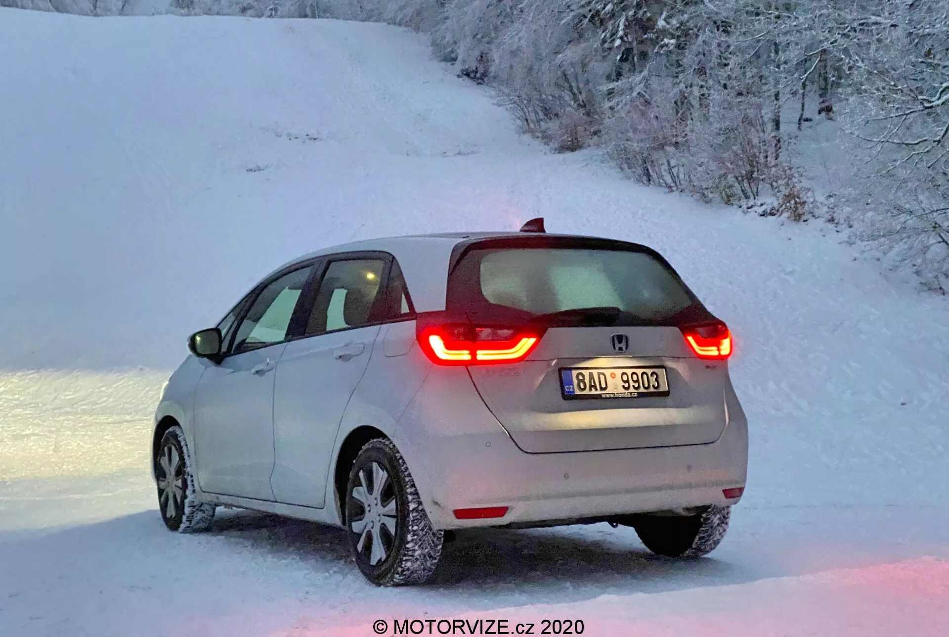 Vista trasera de tres cuartos de un moderno hatchback Honda Jazz 2020, iluminado por la suave luz del amanecer o el atardecer, con luces traseras LED iluminadas en forma de C, un logo del fabricante visible, una ligera capa de nieve en el vehículo y llantas de aleación con neumáticos cubiertos de nieve en un entorno rural nevado.