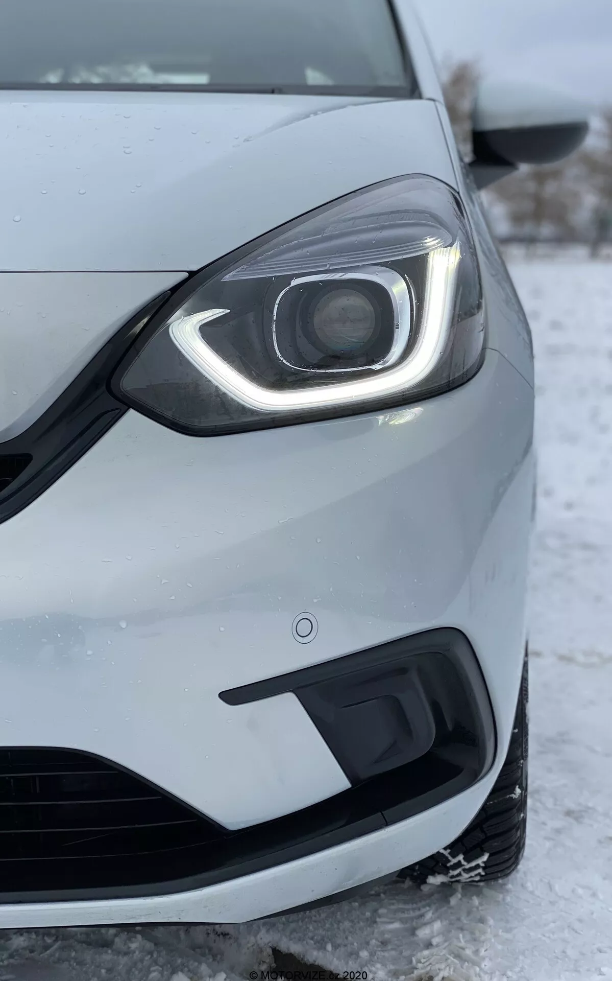 Imagen en primer plano del lado delantero izquierdo de un Honda Jazz 2020, tomada desde un ángulo bajo. El enfoque está en el faro que presenta un diseño moderno con luces de circulación diurna LED que curvan alrededor de la parte superior y lateral. El conjunto del faro incluye una lente de proyector visible, lo que sugiere faros HID o LED avanzados para el haz principal. El parachoques delantero, mostrado debajo del faro, incluye una rejilla de entrada de aire y una carcasa para luces antiniebla. La pintura metálica plateada o gris claro del coche y la rueda parcialmente visible y el espejo lateral están detallados. La nieve y el hielo en el suelo indican un clima frío, lo que refleja el rendimiento del coche en condiciones invernales. La imagen también resalta la calidad de construcción del coche, las brechas entre paneles y el sensor de proximidad en el parachoques, que forma parte de la tecnología de asistencia al aparcamiento.