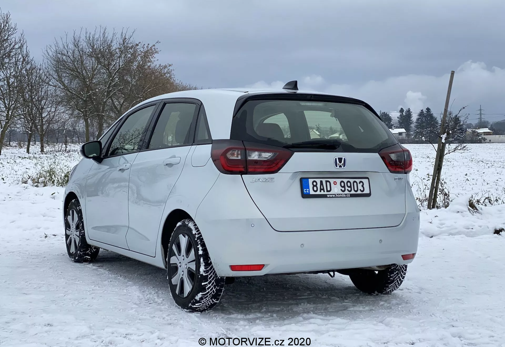 Imagen de un Honda Jazz 2020 plateado estacionado en condiciones nevadas con un paisaje rural o semi-rural en el fondo. Capturado desde la parte trasera y un perfil lateral parcial del lado derecho, destacando las luces traseras dispuestas verticalmente, la insignia central y el parachoques con sensores de aparcamiento. Se aprecian claramente los contornos laterales lisos, las llantas de aleación y los neumáticos de invierno, junto con la capacidad de desempañado en ventanas y parabrisas trasero, y los espejos laterales no plegables del mismo color que la carrocería. También se observa la matrícula, la antena en el techo y el buen estado general del vehículo.