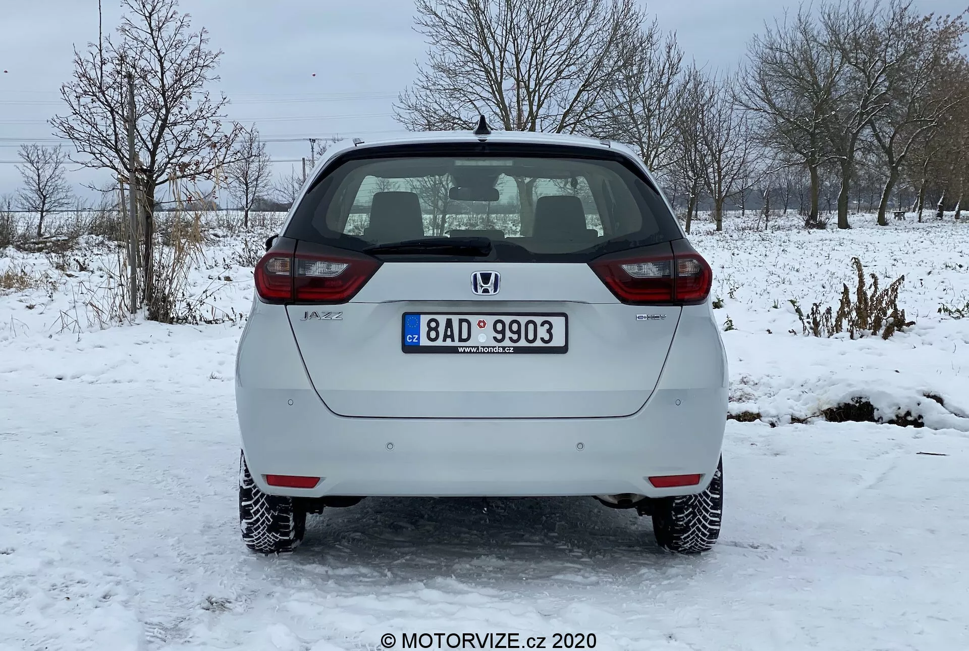 Vista trasera de un coche plateado Honda Jazz 2020 hatchback estacionado sobre una superficie helada. La imagen captura claramente el parachoques trasero, el portón trasero, las luces traseras y el parabrisas trasero del vehículo. El emblema del fabricante y el nombre del modelo son visibles en el portón trasero, junto con una placa de matrícula estándar montada en el centro. El coche cuenta con luces de matrícula iluminadas, reflectores en ambos extremos del parachoques trasero y luces traseras de bombilla estándar. El parabrisas trasero tiene una función de descongelación mostrada por líneas horizontales y un limpiaparabrisas trasero aparcado en la parte superior. El sistema de escape es ligeramente visible debajo del parachoques trasero en el lado derecho. El coche tiene llantas de acero con cubiertas decorativas y está ubicado en un paisaje invernal con nieve y árboles desnudos en el fondo.