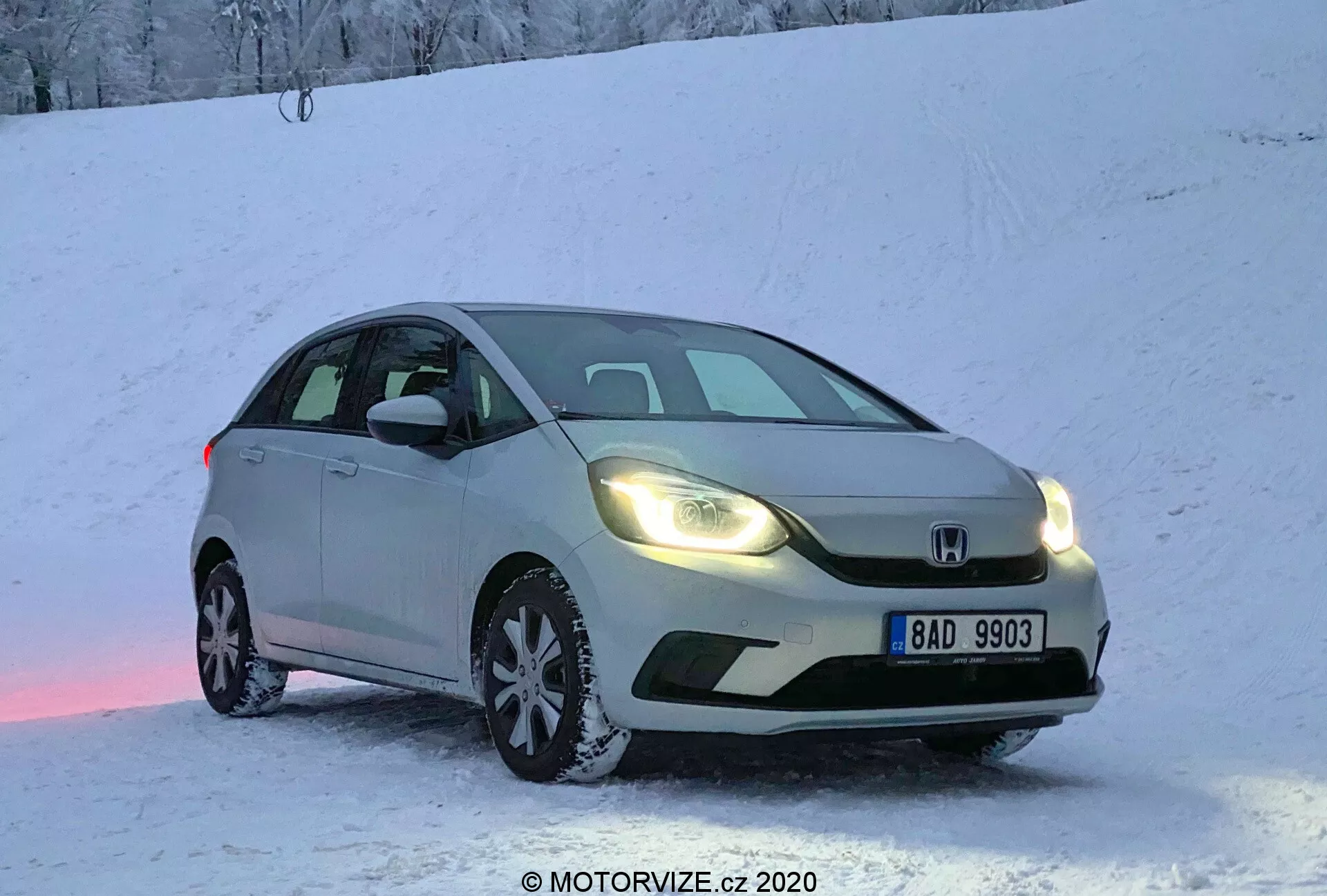 Coche hatchback Honda Jazz 2020 de color plateado estacionado sobre una superficie nevada durante el amanecer o atardecer, con los faros cálidos encendidos, capturado desde una vista tres cuartos frontal. La imagen resalta la parrilla delantera, los faros, el capó, el parabrisas y parte del lado del conductor. Matrícula europea visible, nieve en el suelo y el coche, creando una ambiente fría pero acogedora. Reflexión de luz rosada en la nieve debajo del coche. Toma en ángulo bajo que enfatiza el equipo de neumáticos de invierno y la apariencia dinámica para capacidades de conducción en invierno.