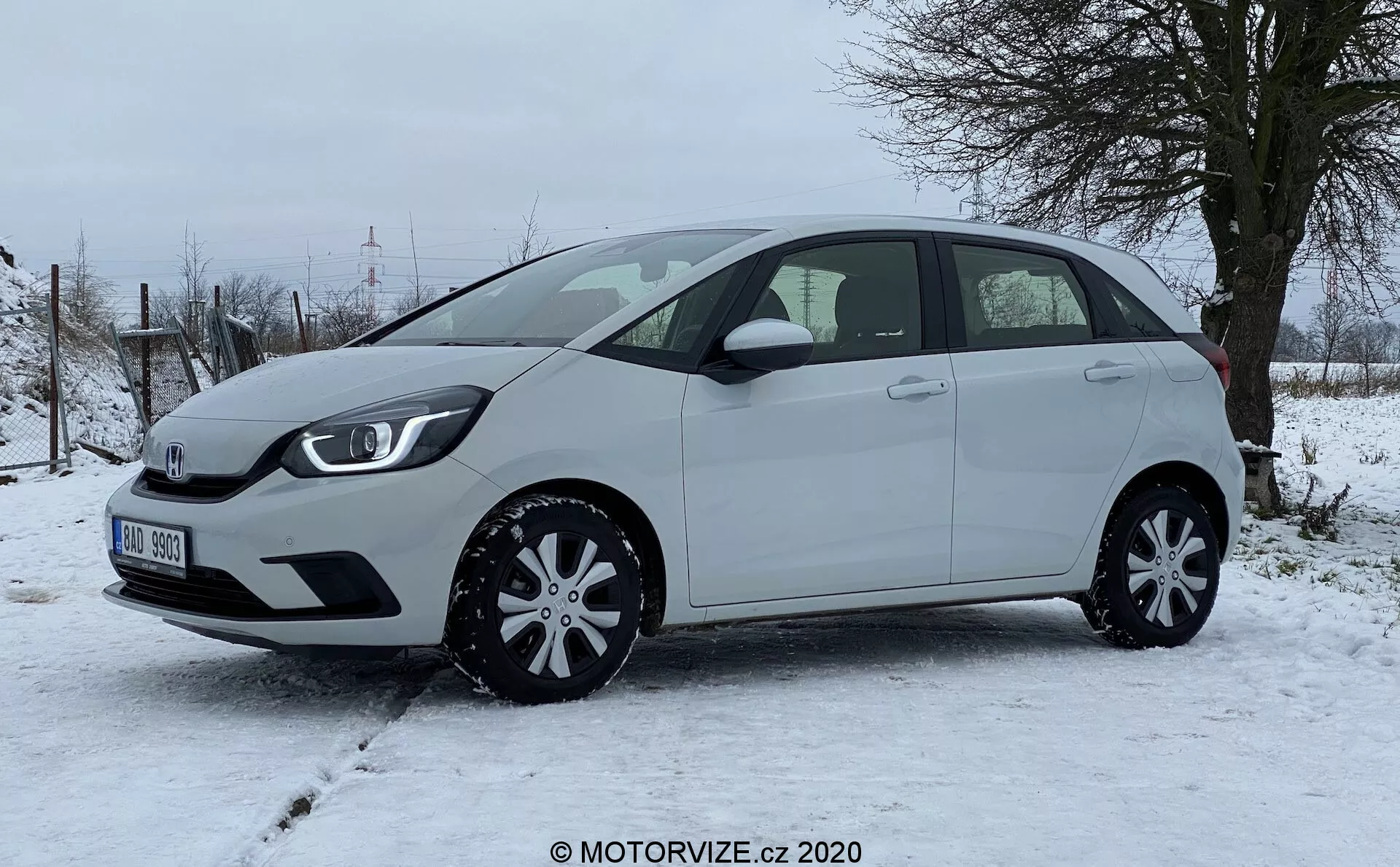 Un deportivo Honda Jazz 2020 hatchback fotografiado de lado, destacando el perfil izquierdo del vehículo en un terreno nevado con vegetación rural en el fondo. El coche compacto presenta una línea de techo ligeramente inclinada, cinco puertas incluyendo una trasera, espejos laterales del color de la carrocería y cubiertas de ruedas elegantes, exudando practicidad y eficiencia en el diseño. No se observan características exteriores avanzadas como cámaras o sensores desde este ángulo, enfatizando una vista clara de las proporciones del coche, contornos de las puertas, línea de las ventanas y arcos de las ruedas.