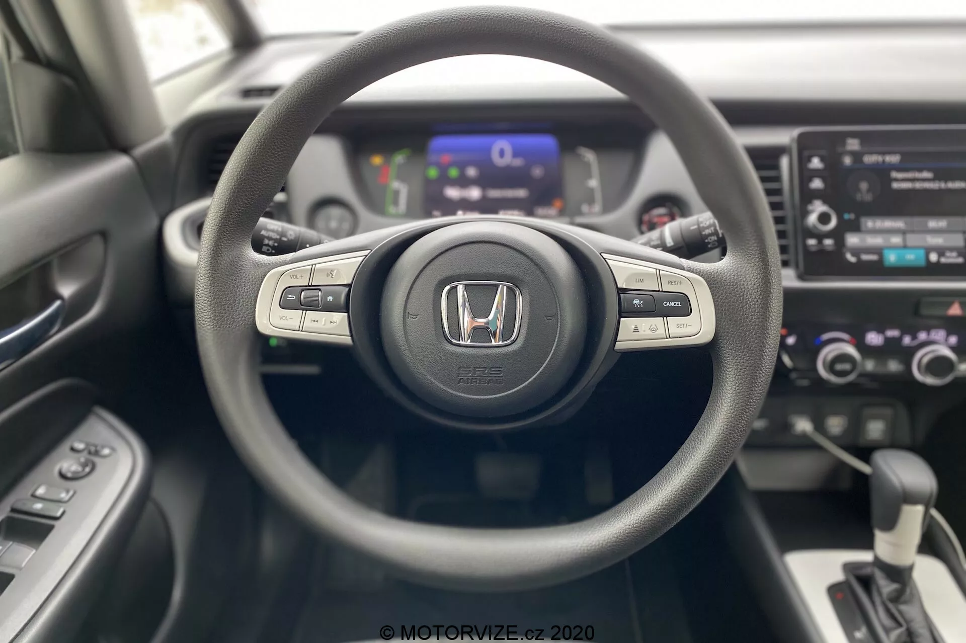 Vista interior del Honda Jazz 2020 desde la perspectiva del conductor, destacando el volante de tres radios con emblema de Honda, botones de control para el sistema de medios y control de crucero en los radios, un panel de instrumentos digital en el fondo ligeramente desenfocado y una consola central con sistema de infoentretenimiento táctil, controles de climatización de doble zona y palanca de cambios automática. Diseño moderno y contemporáneo con materiales de texturas mixtas.