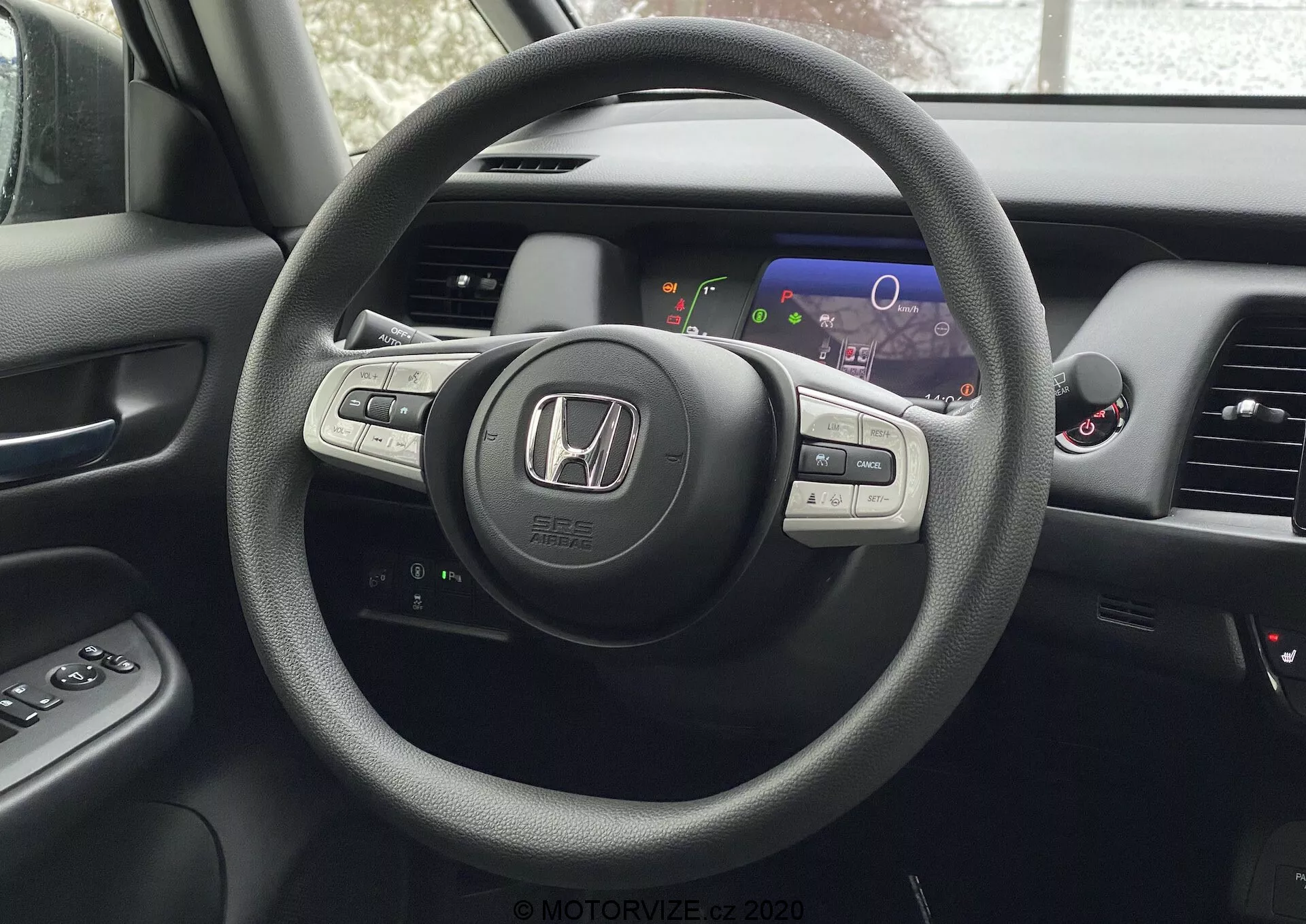 Vista interior de un Honda Jazz 2020 desde la perspectiva del asiento del conductor, destacando el volante de tres radios con el emblema de Honda y controles montados para el audio, control de crucero y probablemente Bluetooth o control por voz. Detrás del volante hay un panel de instrumentos digital con pantallas coloridas que incluyen un velocímetro y un cuentavueltas. Parcialmente visible está la consola central con pantalla de información y entretenimiento o navegación y las rejillas de ventilación. También es parcialmente visible el panel de la puerta del conductor con botones de control de ventanas y espejos laterales.