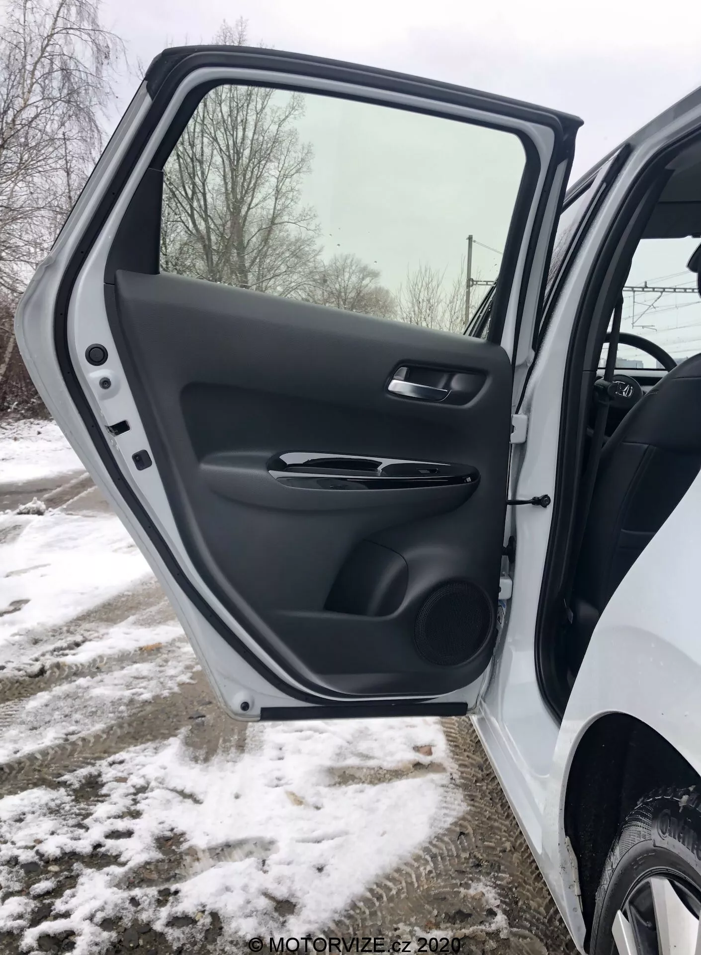 Primer plano del interior de la puerta delantera del lado del conductor del Honda Jazz 2020, mostrando una vista detallada del acabado plástico texturizado, la manija ergonómica satinada, controles de la ventana y espejo eléctrico, bolsillo de almacenamiento moldeado y rejilla del altavoz, todo ambientado en un entorno invernal nevado.
