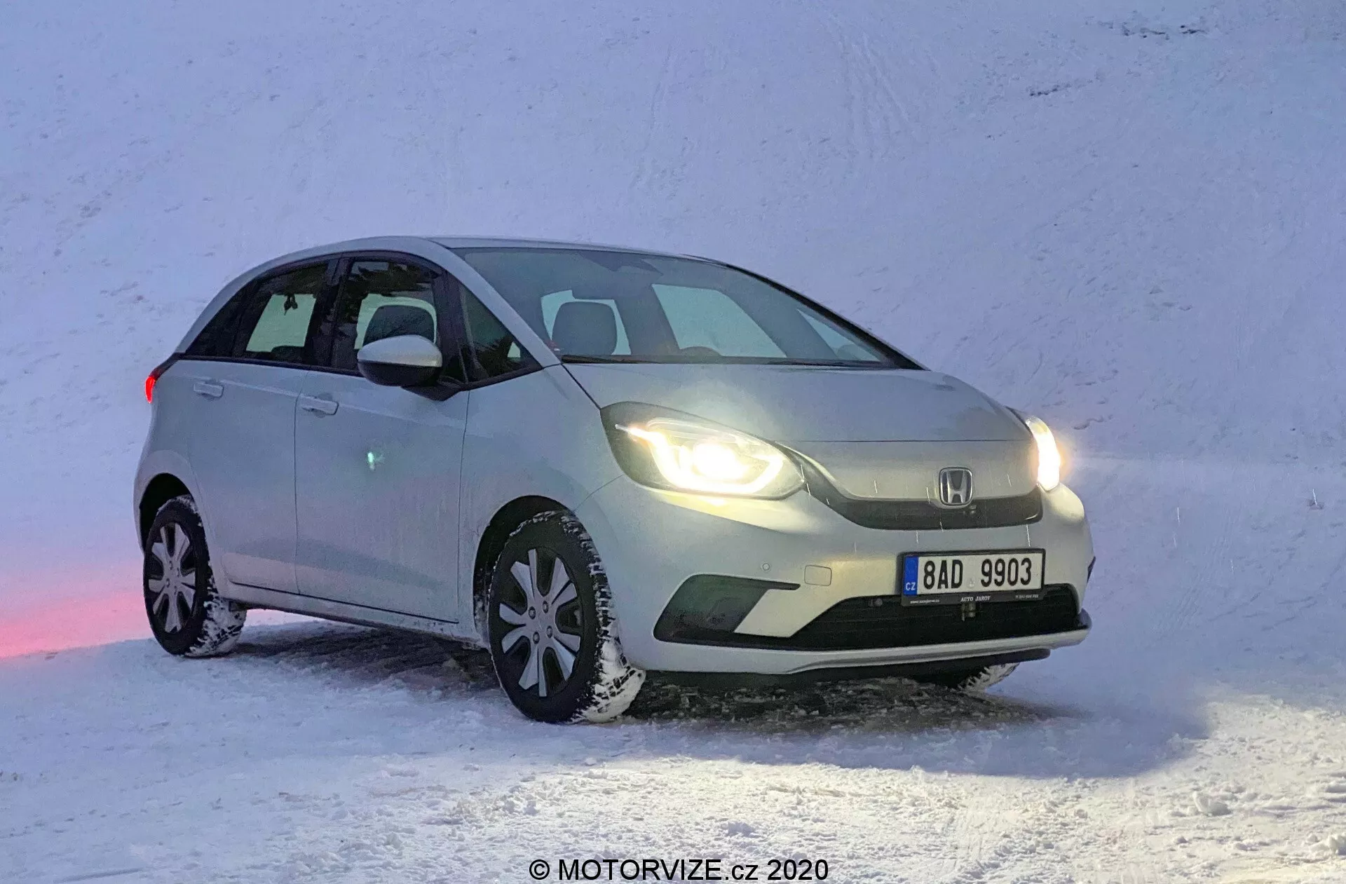 Una vista frontal de tres cuartos de un hatchback Honda Jazz 2020 de color claro, posicionado sobre una superficie nevada. La imagen captura el coche desde el lado delantero izquierdo, mostrando el lado del conductor en un ángulo que resalta tanto el perfil lateral como la fachada frontal. El Honda Jazz está estacionado con el frente ligeramente hacia la derecha, revelando el diseño de la parrilla, los faros y parte del capó. Los faros delanteros están encendidos, reflejándose en la nieve, con el vehículo aparentemente parado. Los neumáticos de nieve con patrones de rodadura distintivos son visibles, destacando su idoneidad para la conducción en invierno.
