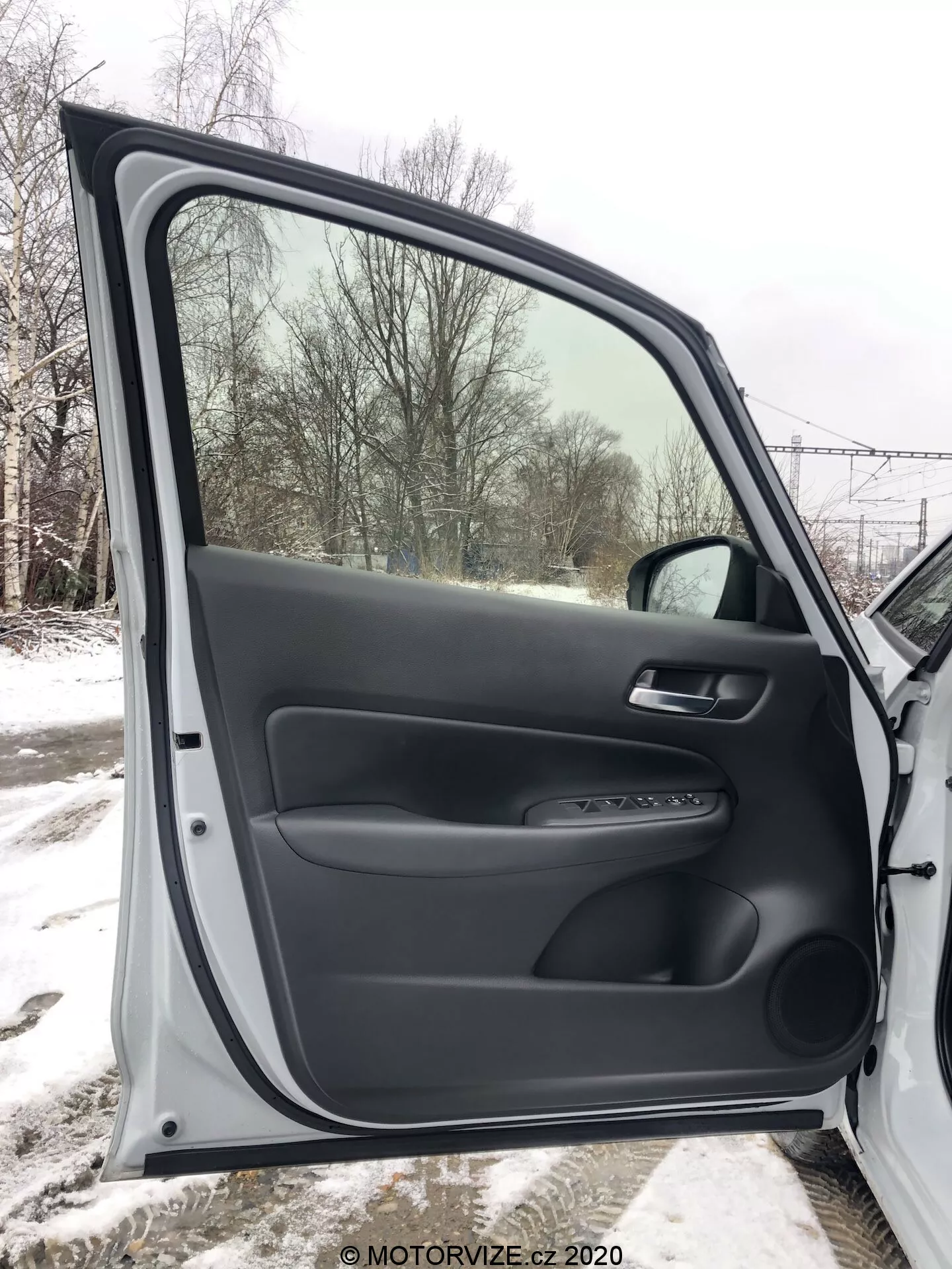Vista interior de la puerta delantera del pasajero de un Honda Jazz 2020, completamente abierta, presentando materiales plásticos negros tanto texturizados como lisos. El panel de la puerta incluye un reposabrazos ergonómico, una manija de material suave, y un panel de control para los mecanismos de la ventana y cerradura eléctricas. También cuenta con un acento de trim plateado y un bolsillo de almacenamiento inferior. A través de la ventana, se ve un paisaje nevado, indicando un entorno invernal.