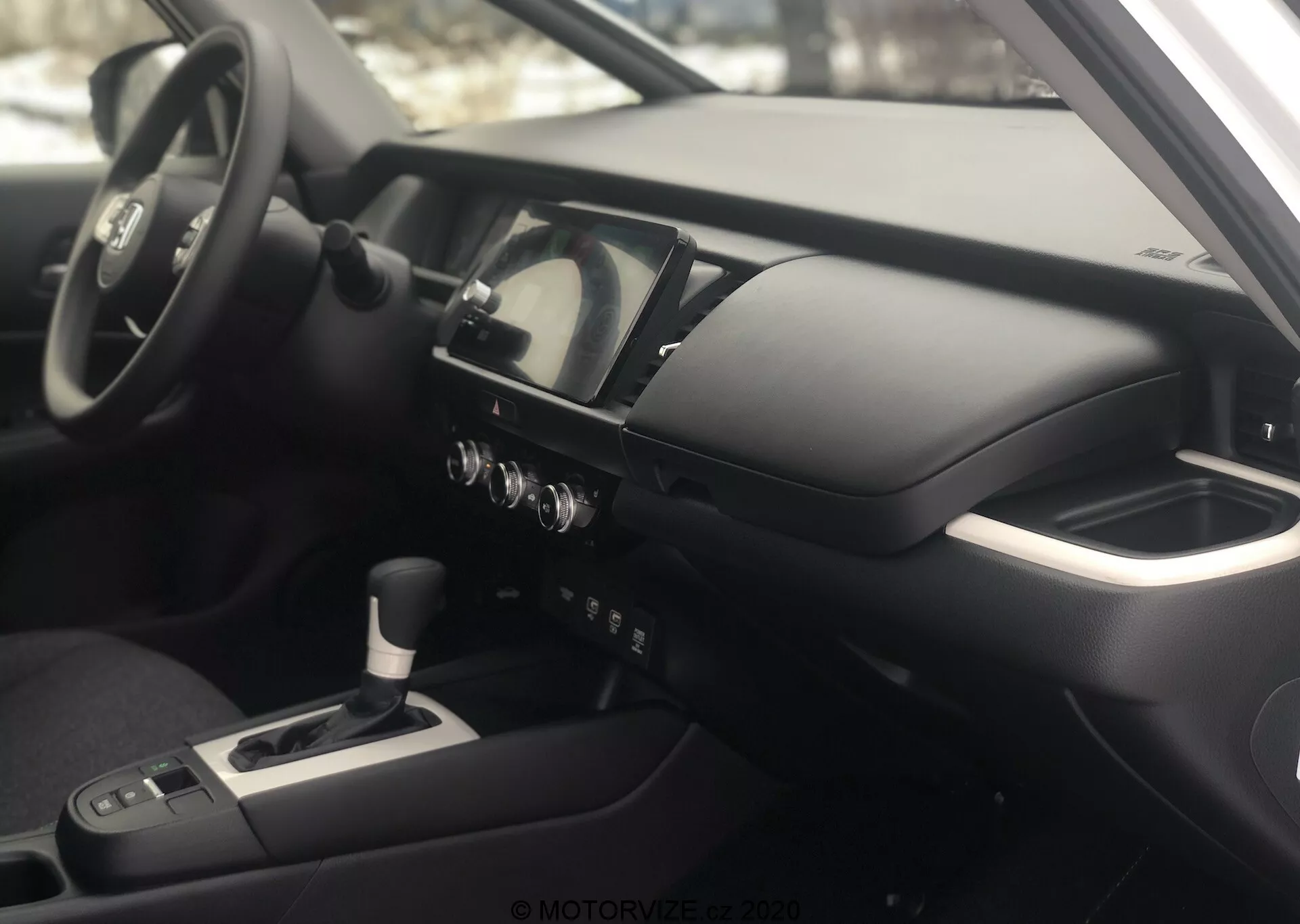 Vista interior del Honda Jazz 2020 desde el lado del pasajero, enfocándose en el tablero y la cabina del conductor. La fotografía, tomada en un ángulo ligeramente superior, resalta la consola central, el volante multifuncional con controles montados, el panel de instrumentos digital-analógico, la consola central con palanca de cambios, los botones de control del clima y una pantalla de infoentretenimiento/navegación. El reposabrazos sugiere comodidad para el conductor, y el interior cuenta con materiales metálicos y suaves al tacto, reflejando una filosofía de diseño moderna y de alta gama.
