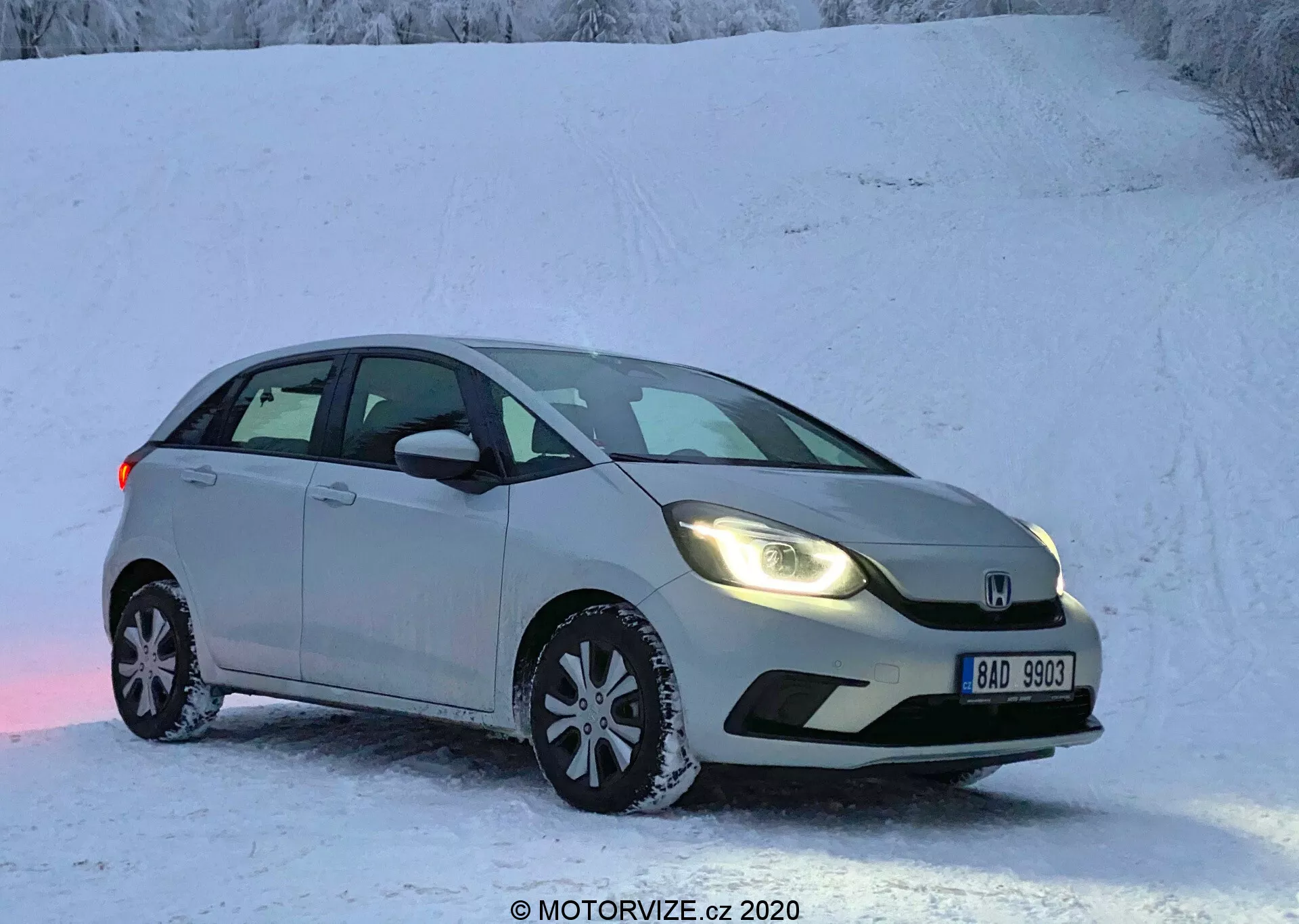 La imagen muestra un Honda Jazz 2020, un hatchback plateado o gris claro estacionado en una superficie nevada, sugerente de una escena invernal con un paisaje cubierto de nieve. Capturado desde un ángulo frontal de tres cuartos, la vista resalta el lado del conductor, mostrando la parrilla delantera, faros, capó, parabrisas, puerta del conductor y puerta trasera del pasajero. El coche cuenta con faros modernos, un parachoques delantero contorneado, una insignia prominente en la parrilla, espejos laterales con señales de giro integradas y ruedas de aleación de múltiples radios equipadas con neumáticos de invierno. El vehículo estacionario se muestra sin desenfoque de movimiento bajo posibles condiciones de poca luz, ya sea temprano en la mañana o al final de la tarde.