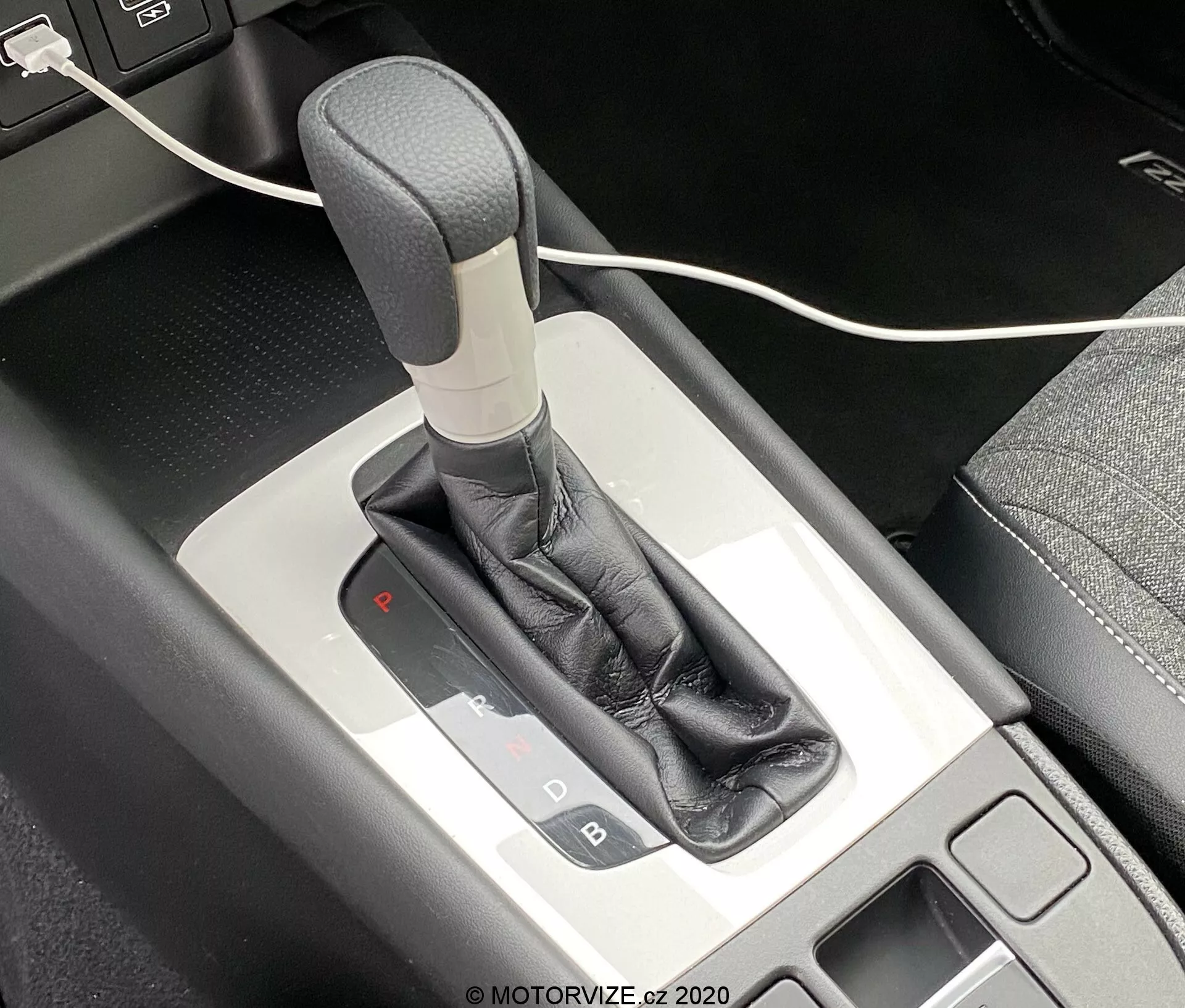 Vista de cerca de la palanca de cambios de transmisión automática del Honda Jazz 2020 desde un ángulo cenital, mostrando el mecanismo del selector de cambios y la consola central circundante. La palanca de cambios tiene un mango gris o plateado, envuelto en una funda de cuero negro, complementado con un borde metálico. El panel de selección de cambios iluminado indica que el vehículo está en modo de estacionamiento con una luz roja debajo de la 'P'. Se puede ver un pequeño área de almacenamiento con una alfombrilla negra a la izquierda, junto con un cable USB parcialmente visible en la parte superior de la imagen.