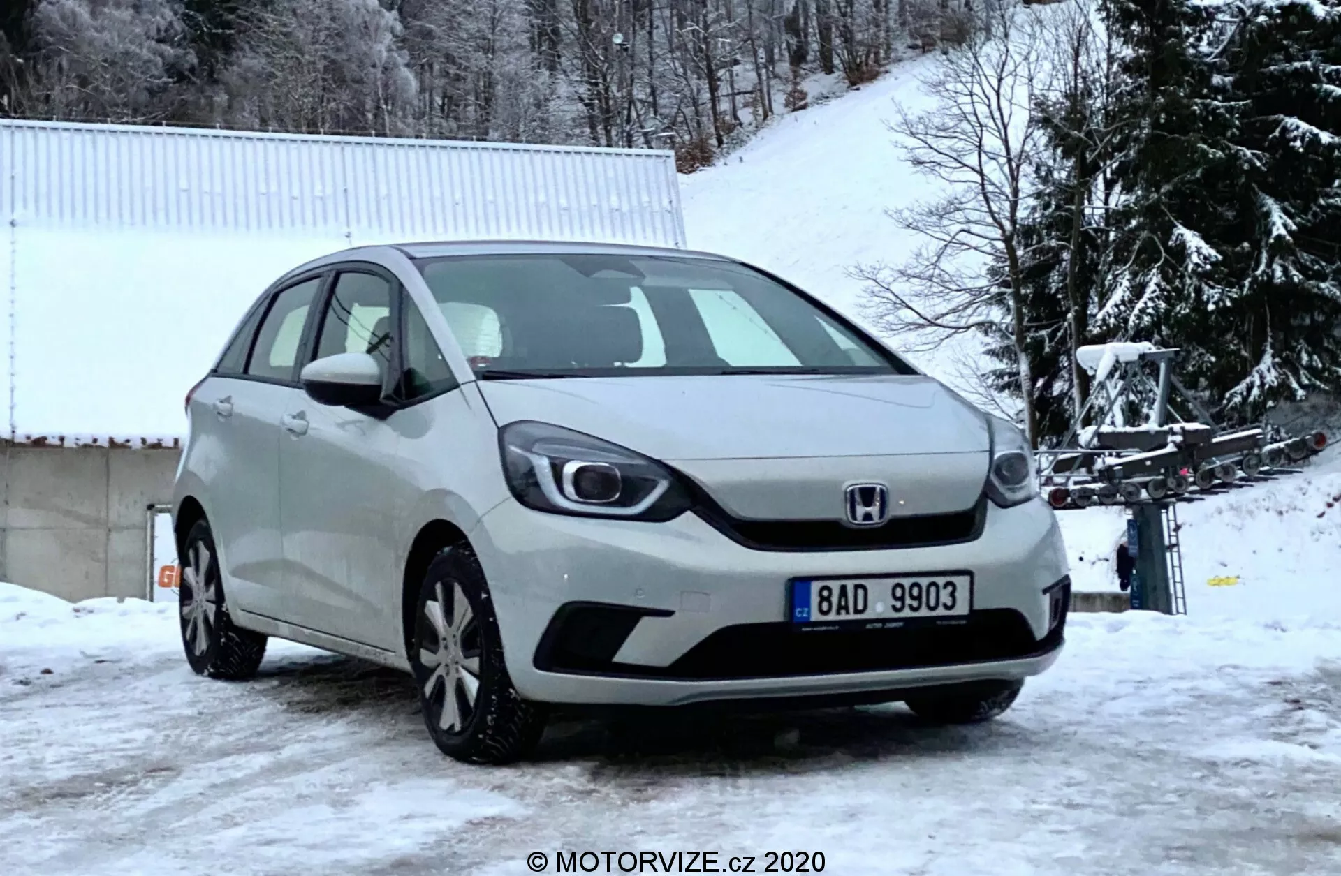 Vista en ángulo frontal de un Honda Jazz plateado 2020 capturado en un día invernal, mostrando la parrilla delantera, faros, capó y parachoques delantero contra un fondo cubierto de nieve con árboles desnudos y un edificio blanco.