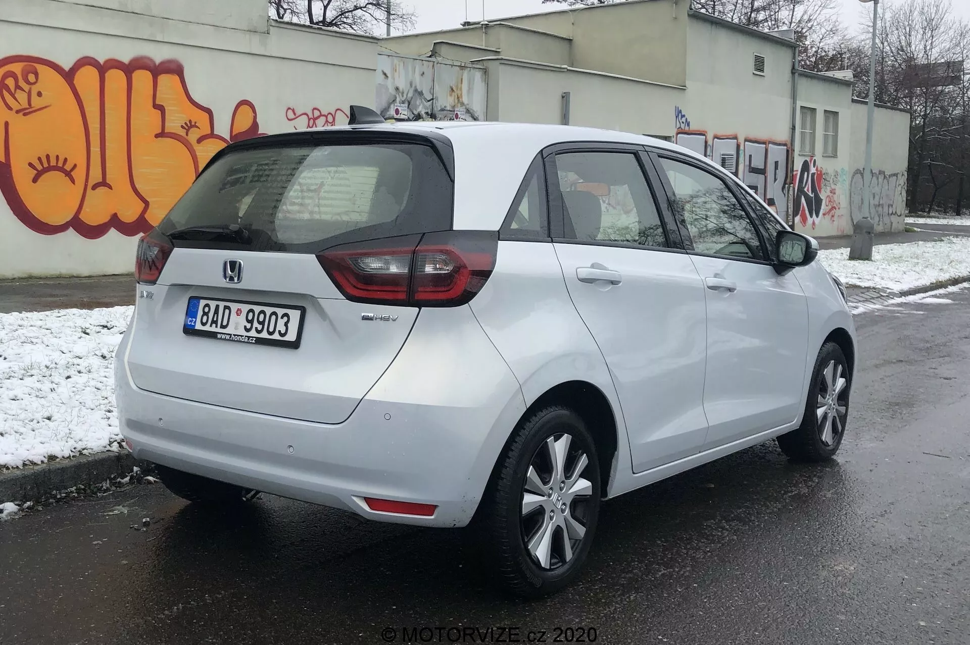 Vista trasera de tres cuartos de un Honda Jazz 2020 blanco estacionado en una superficie de asfalto en un entorno urbano potencialmente frío con nieve en el suelo y graffiti en la pared. El coche presenta una combinación de luces traseras de LED y bombillas tradicionales, placa de matrícula central trasera y una insignia que indica el nivel de acabado o variante. Tiene un techo aerodinámico, llantas de aleación de cinco radios, espejos laterales del color de la carrocería con indicadores de giro integrados y manijas de puerta a juego con el color de la carrocería. El parabrisas trasero tiene un desescarchador y un limpiaparabrisas para una mejor visibilidad.