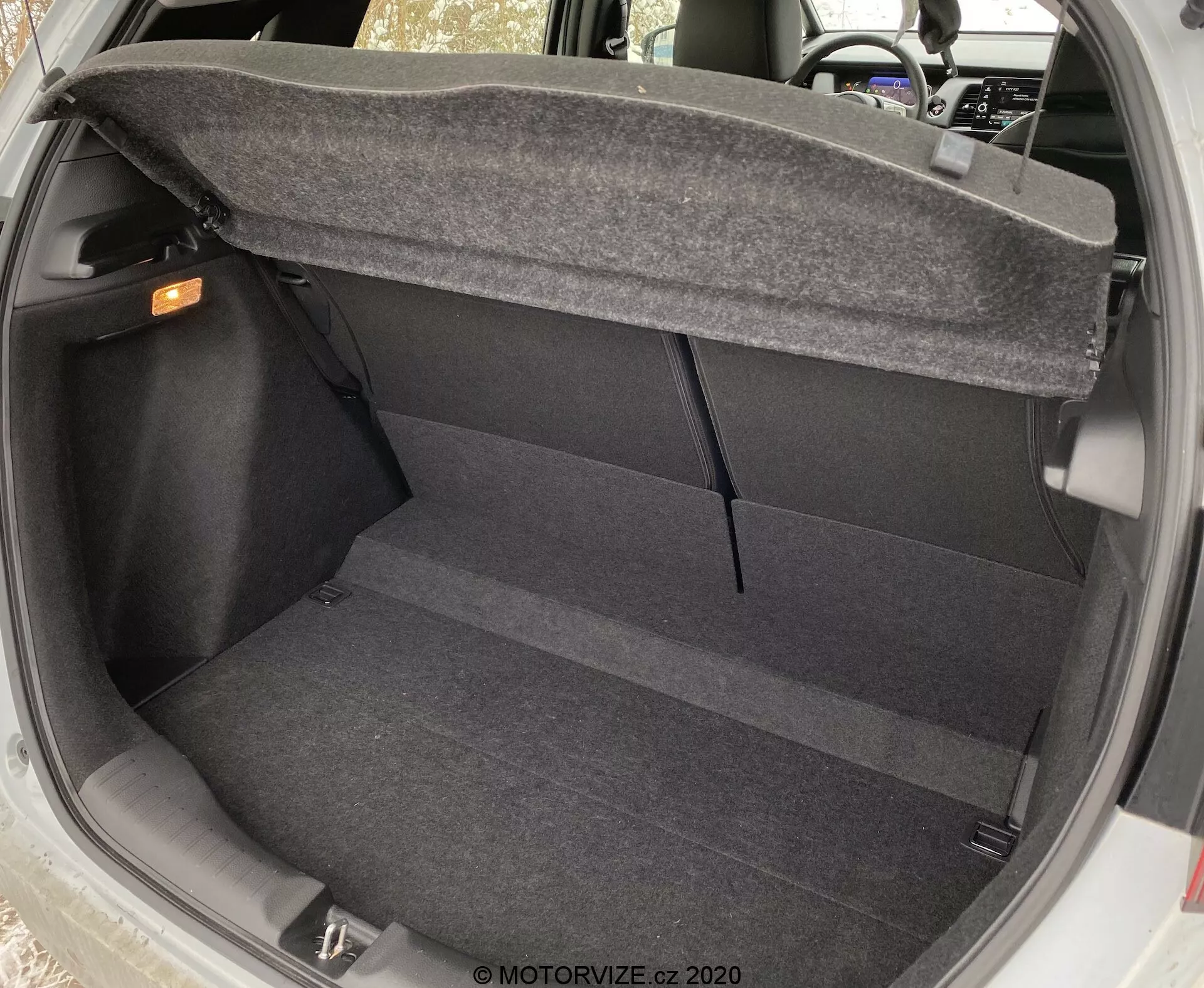 Vista interior del maletero del Honda Jazz 2020 desde un ángulo ligeramente elevado, mostrando el suelo alfombrado limpio, las paredes laterales y la parte trasera de los asientos traseros. El maletero está vacío, con el tirador de liberación interior visible en la esquina superior derecha y el mecanismo de bisagra de la tapa del maletero parcialmente visible en los laterales superiores, destacando la practicidad y las características de diseño básicas del maletero.