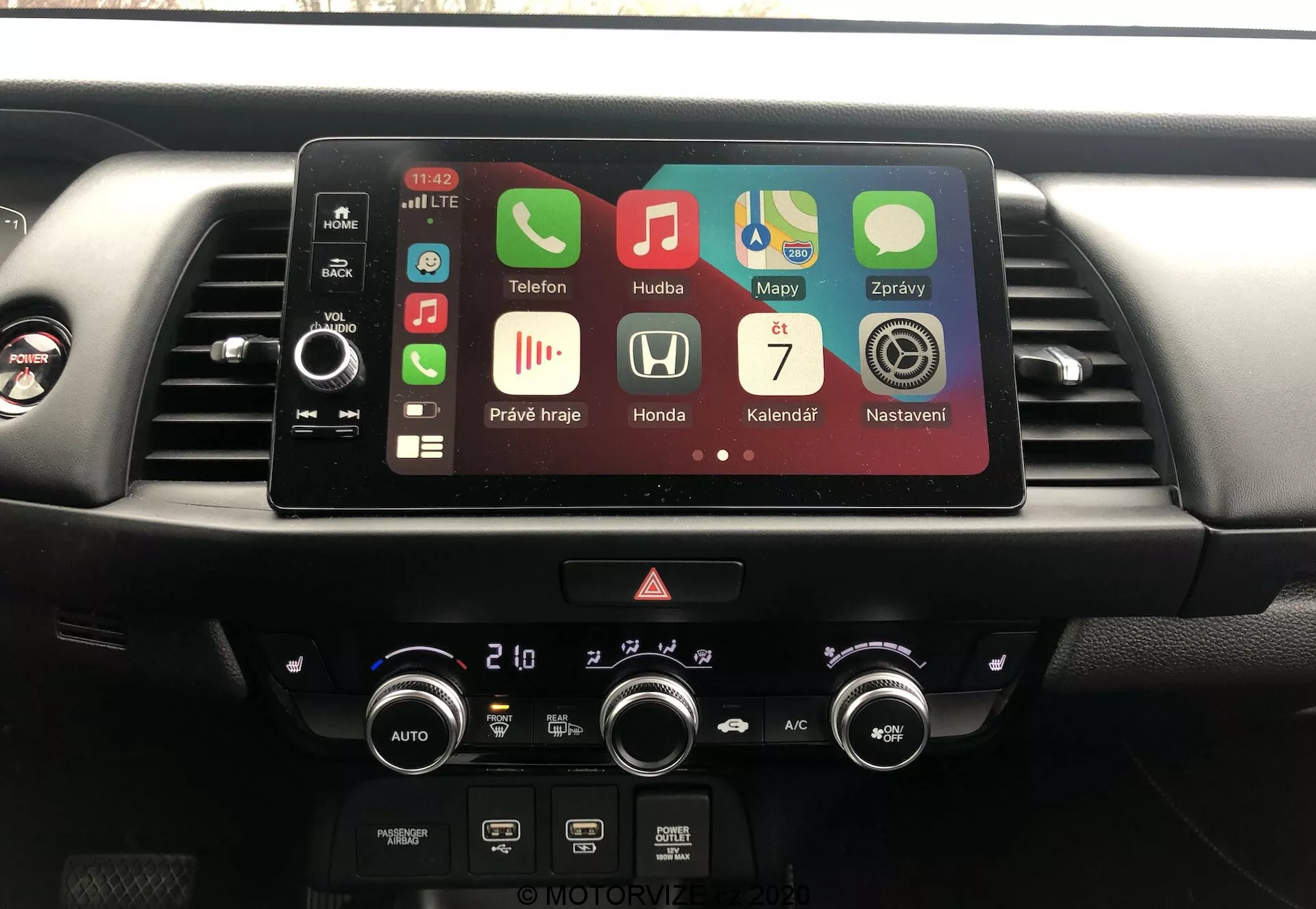 Consola central del interior del Honda Jazz 2020, mostrando sistema de infoentretenimiento y control del clima desde la perspectiva del conductor. Pantalla táctil activa con aplicaciones como Teléfono, Música, Mapas que indican Bluetooth, GPS, reproducción de medios y Apple CarPlay. Debajo de la pantalla, botones físicos, luces de emergencia y diales de control climático de doble zona con lecturas en Celsius.