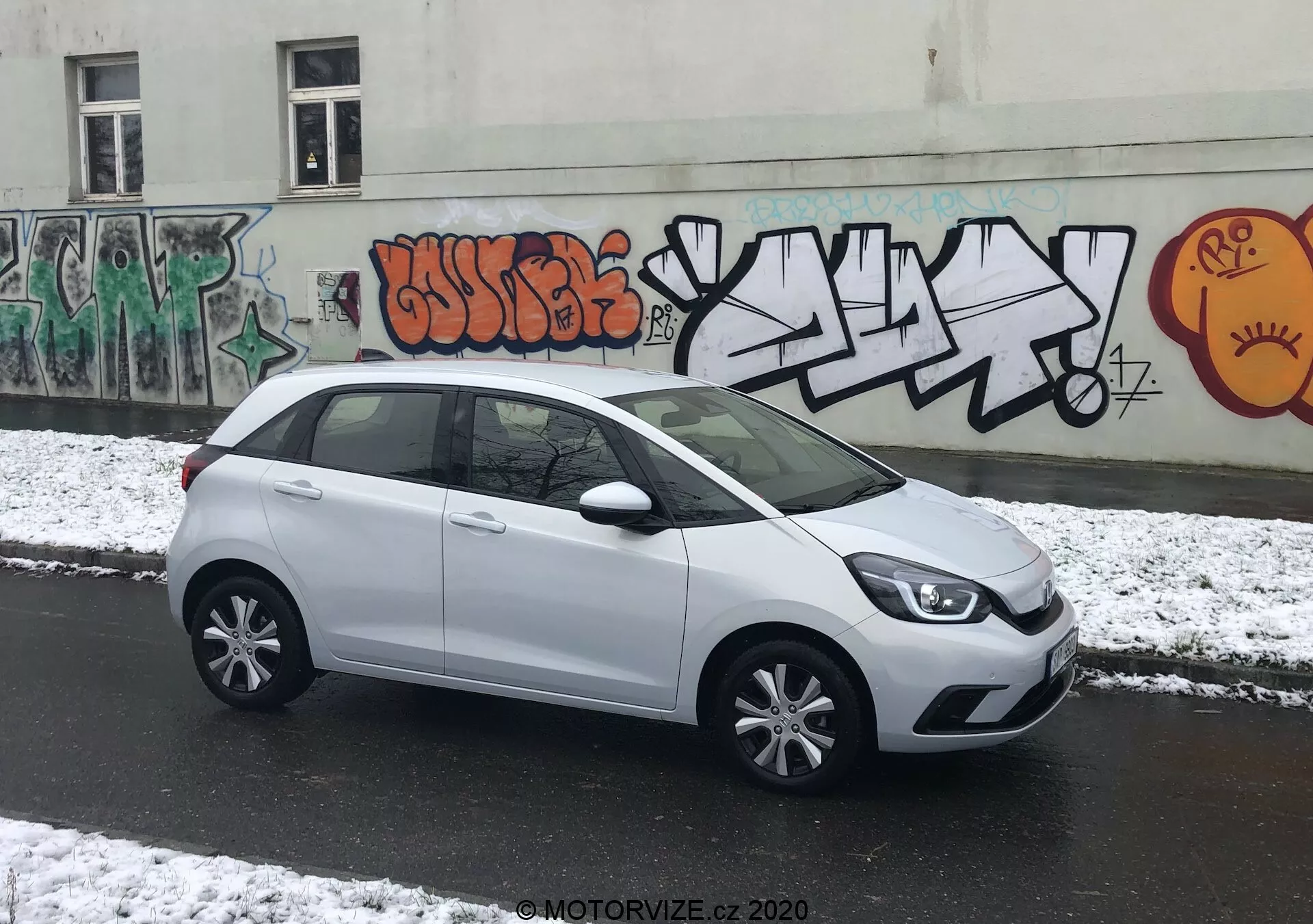 La imagen muestra un hatchback Honda Jazz 2020 blanco estacionado perpendicularmente a la cámara en una calle urbana, permitiendo una vista completa de su perfil lateral. La parte delantera estilizada del coche con modernos grupos de faros es parcialmente visible, conduciendo hacia una suave línea de carácter en el costado. Las manijas de las puertas, espejos laterales a juego con el color del cuerpo y las cubiertas de rines de múltiples radios realzan su apariencia elegante. La amplia zona acristalada ofrece amplio espacio para la cabeza, y una ventana trasera pequeña añade a su diseño. El spoiler trasero sugiere eficiencia aerodinámica. La iluminación nublada con nieve fresca y el telón de fondo urbano con graffiti añade un entorno dinámico.