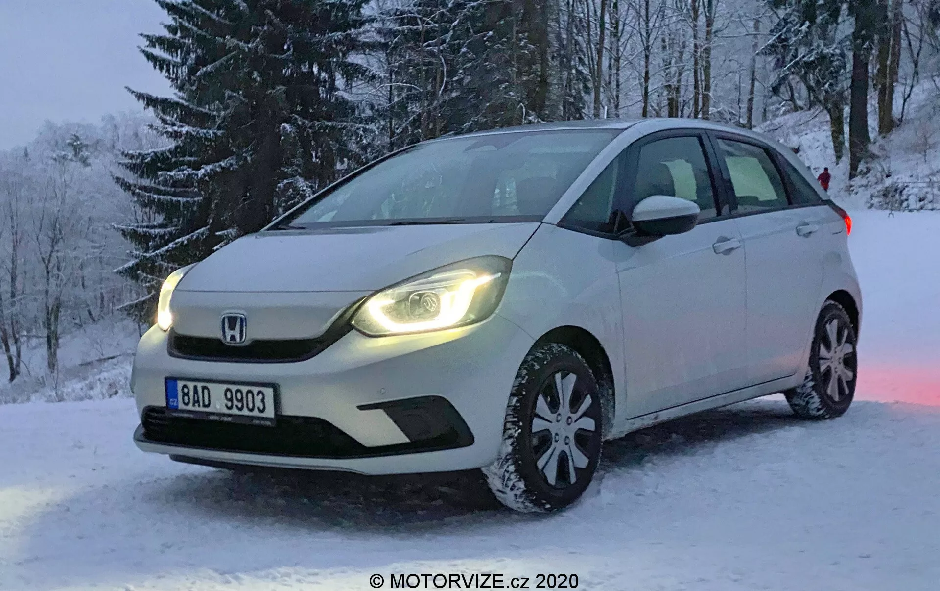 Fotografía profesional de un hatchback Honda Jazz blanco del 2020 estacionado en un terreno nevado durante el invierno. La imagen se toma en un ángulo que enfoca el lado frontal izquierdo del automóvil, mostrando la rejilla frontal, los faros LED, el capó, la llanta de aleación multirradio equipada con neumáticos para nieve, el parabrisas y la puerta del conductor. En el fondo se pueden ver árboles cubiertos de nieve y la matrícula es legible. La luz tenue sugiere la mañana temprano o la tarde, con las luces diurnas del automóvil reflejándose en la nieve, realzando la escena. El vehículo presenta iluminación avanzada y señales de giro integradas en los espejos laterales, con el distintivo emblema de Honda en la rejilla prominentemente exhibido.
