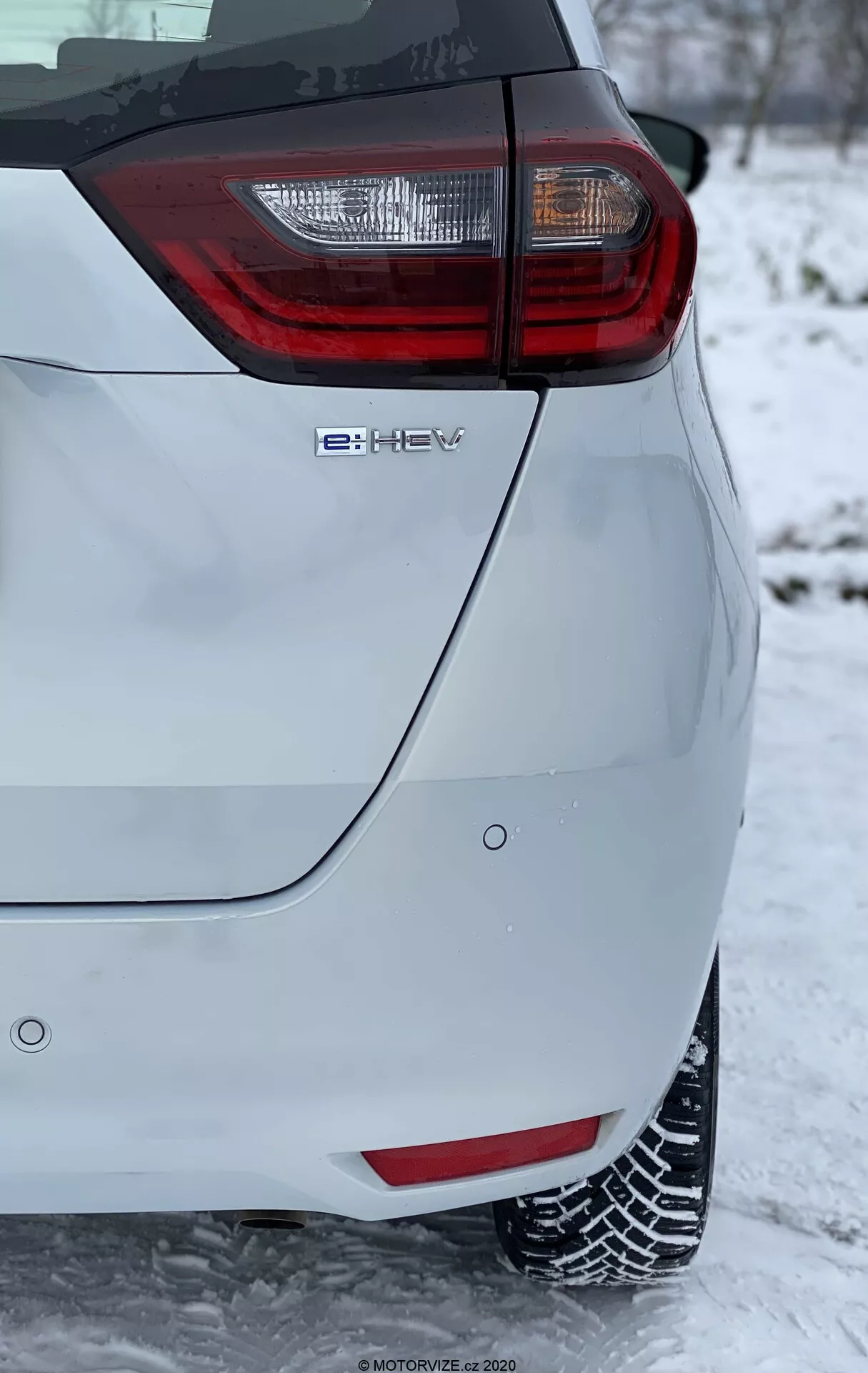 Primer plano de la esquina trasera de un coche hatchback moderno, que presenta un conjunto de luces traseras prominentes con elementos rojos y claros. La imagen, tomada en orientación vertical, muestra el emblema de tecnología híbrida 'e-HEV' y está ambientada en un entorno invernal con nieve en el suelo y cubriendo los neumáticos, indicando condiciones de conducción en invierno. La pintura de color claro del vehículo contrasta con el fondo nevado, destacando sus características de diseño moderno y ecológico.