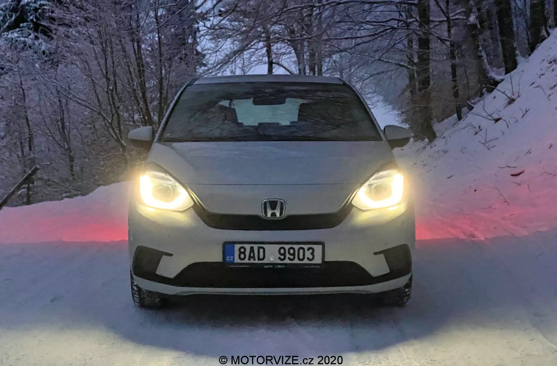 Toma frontal de un Honda Jazz 2020 iluminado con faros LED, mostrando el emblema de la marca y la placa en la parrilla, con un capó esculpido y parabrisas tintado. El suelo nevado y la atmósfera crepuscular realzan el diseño avanzado y la capacidad de conducción invernal del automóvil.