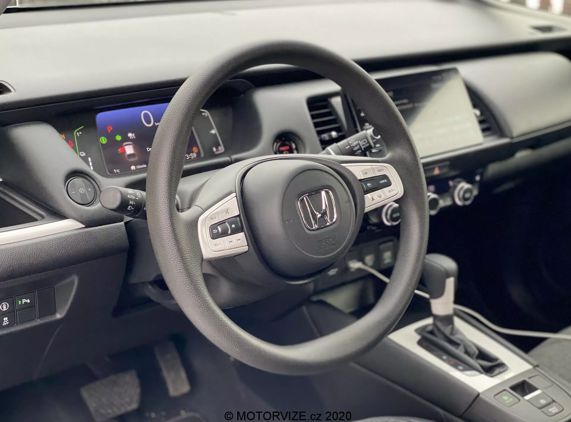 La foto muestra el interior de un Honda Jazz 2020 desde la perspectiva del conductor. Se enfoca en el volante con la marca Honda, equipado con varios botones de control que sugieren funcionalidad integrada de audio, pantalla de información y posiblemente control de crucero. Detrás del volante, se ve una pantalla digital parcial que muestra un indicador de nivel de batería, insinuando que el vehículo podría ser híbrido o completamente eléctrico. El interior bien iluminado y moderno también incluye una consola central con un sistema de infoentretenimiento táctil, controles de clima y una palanca de cambios de diseño contemporáneo con transmisión automática. La imagen resalta los acabados mate, satinado y brillo utilizados en los materiales del interior del coche, enfatizando un diseño ergonómico y rico en tecnología.