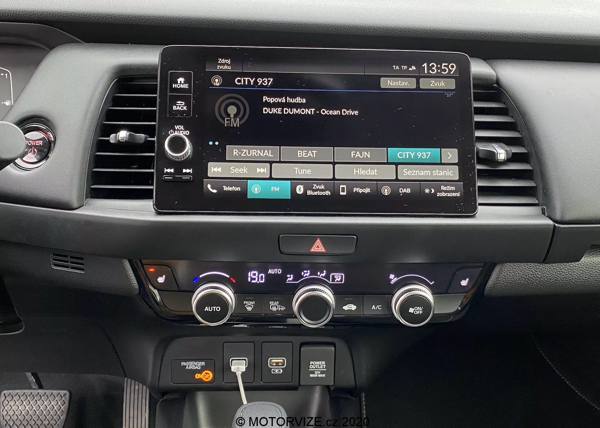 Vista en primer plano del área del tablero central del Honda Jazz 2020. La imagen presenta destacadamente la pantalla táctil del sistema de infoentretenimiento, rodeada de botones y perillas físicas para ajustes de audio, medios y del vehículo. Debajo hay dos ventilaciones de aire y un sistema de control climático con botones y perillas para ajustar la temperatura, velocidad del ventilador, funciones de desempañado y recirculación. El tablero está compuesto por materiales modernos, incluyendo plásticos mate y brillantes, diseñado con accesibilidad funcional y facilidad de uso en mente.