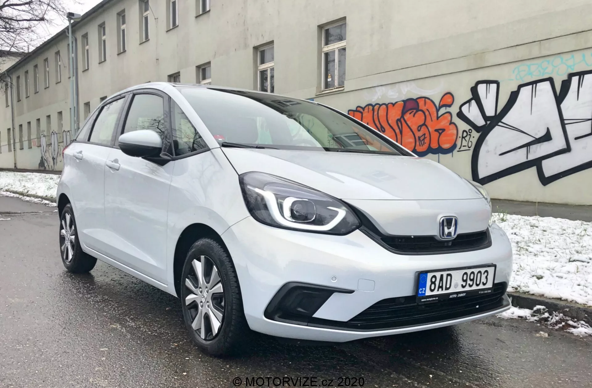 La imagen muestra un vehículo hatchback Honda Jazz 2020 blanco desde un ángulo de tres cuartos desde el frente, revelando sus faros elegantes, la característica parrilla de Honda con un diseño de dos barras y las líneas de contorno aerodinámicas en el capó. El parachoques delantero presenta entradas de aire visibles y posiblemente luces antiniebla, mientras que el perfil lateral muestra ruedas de aleación de múltiples radios y paneles de puerta ligeramente contorneados. El coche está estacionado en una calle nevada, indicando un entorno invernal, con una pared gris decorada con graffiti en el fondo, destacando un entorno urbano.