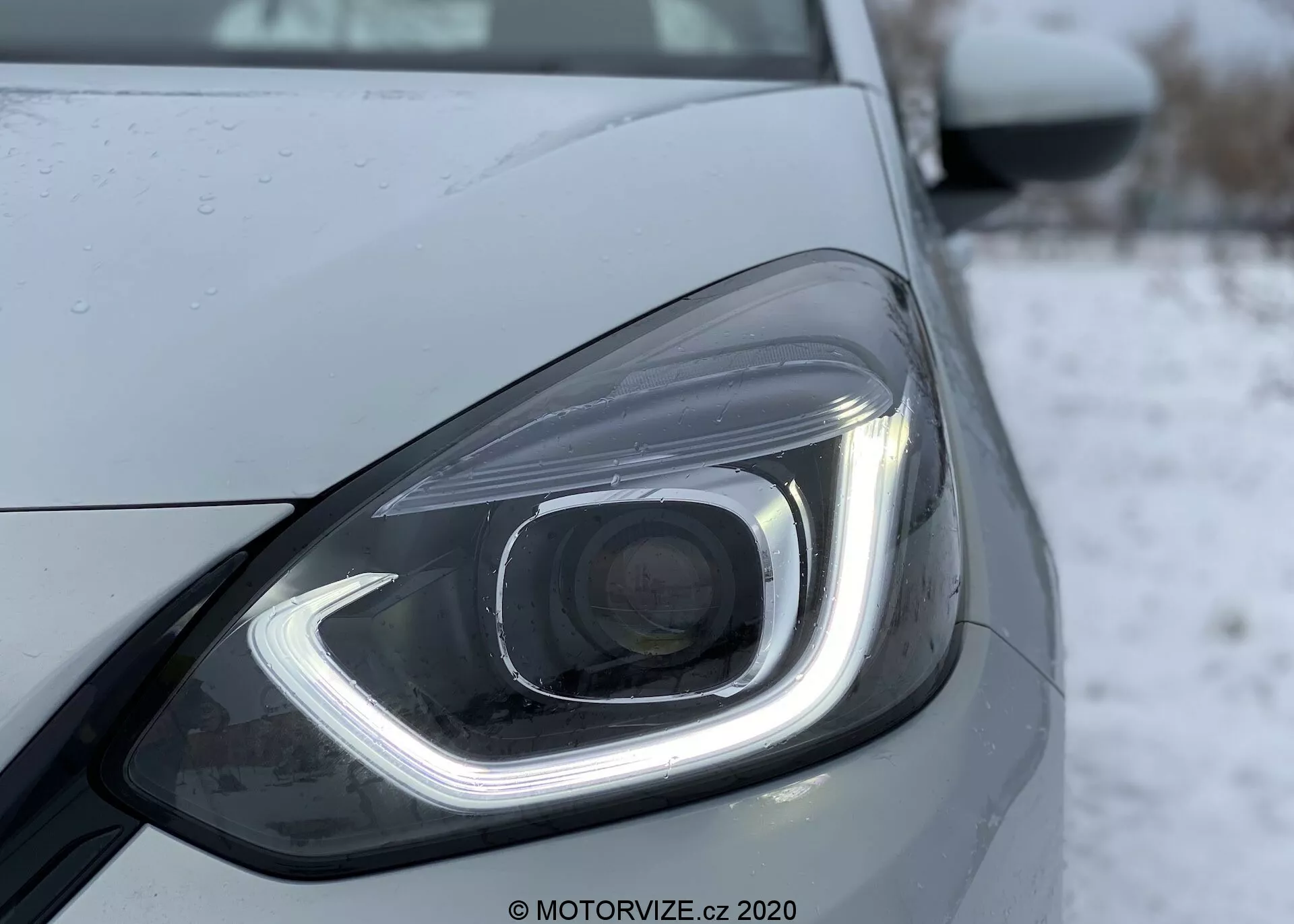 Fotografía en primer plano del faro delantero izquierdo de un Honda Jazz 2020, que muestra su diseño moderno con elementos de iluminación LED o HID. El faro presenta una luz diurna distintiva en forma de gancho, una lente de proyector y gotas de agua que sugieren precipitación reciente o lavado. El diseño elegante se enfatiza con líneas y contornos suaves, con un fondo frío o invernal posiblemente nevado.